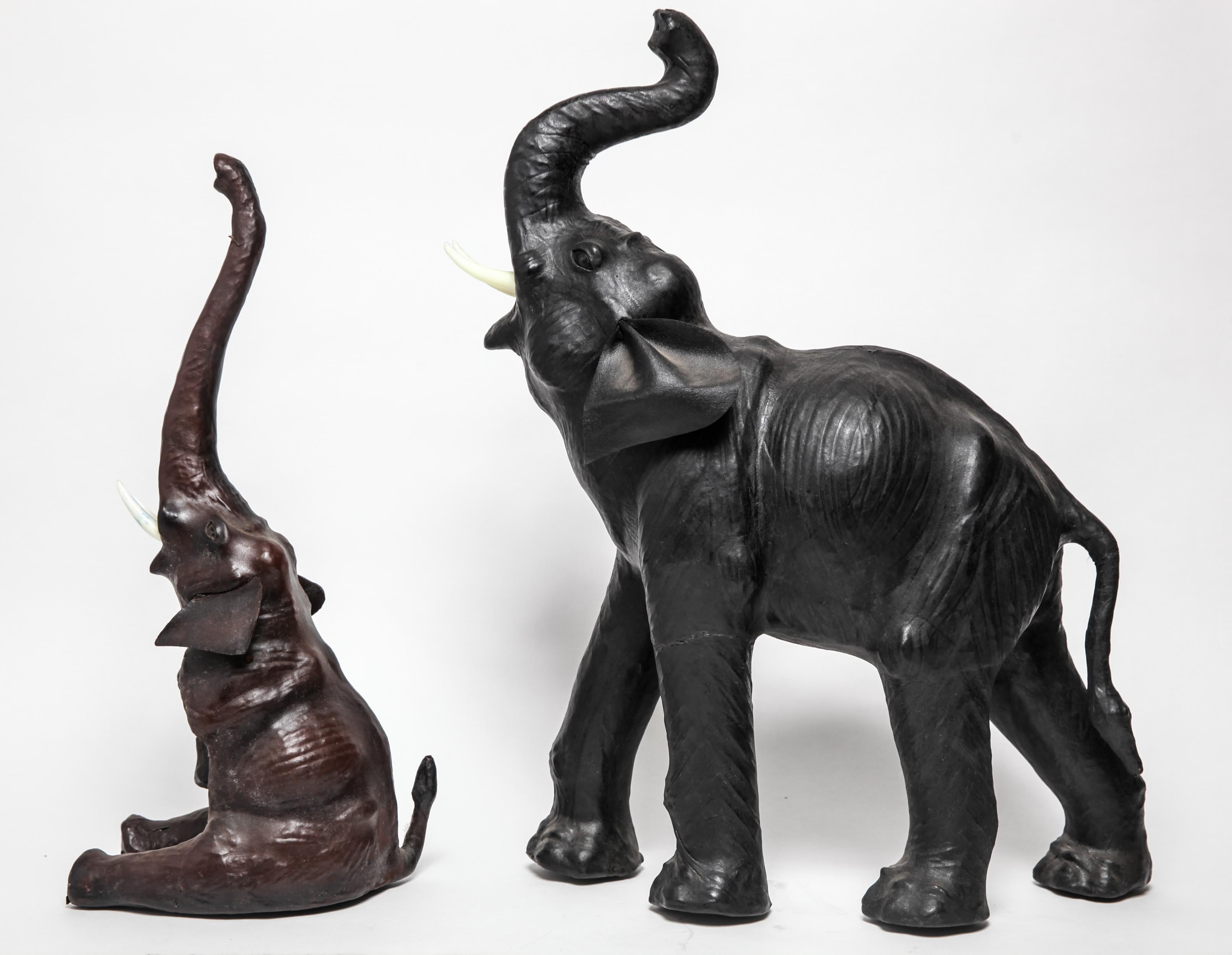 Modern pair of elephant sculptures, one standing and the other seated, both leather clad, with glass eyes and white plastic tusks. The pair is in great vintage condition with some minimal age-appropriate wear to the bottoms.