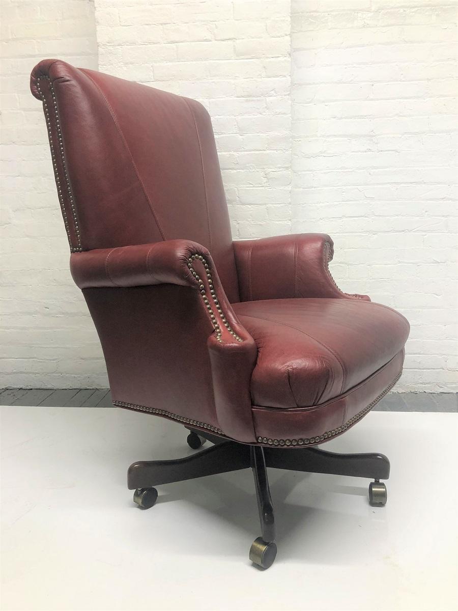 Modern, burgundy leather office chair. The chair has a wooden base with casters, brass studded trim and the chair also swivels.