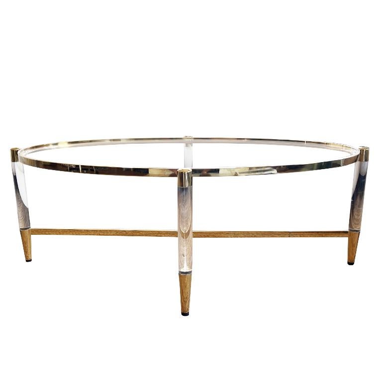 A gorgeous contemporary lucite and brass glass coffee or cocktail table. This piece will be the focal point in any living room or formal sitting area. It is oval in shape and has a brass X base. Four transparent acrylic or lucite legs are affixed on