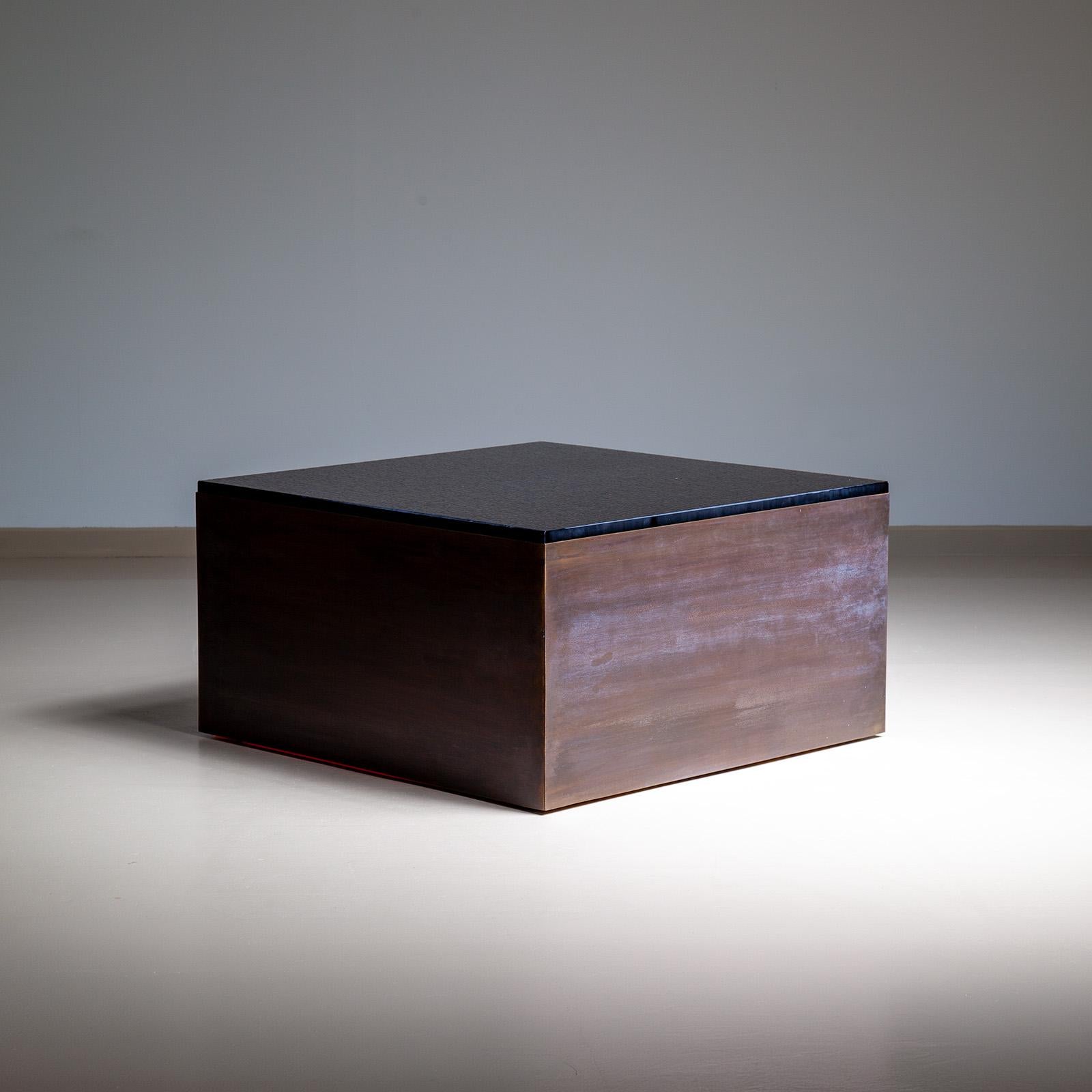 Modern gestalteter Couchtisch mit einem quadratischen Sockel aus gebürstetem Metall und einer dicken, strukturierten braunen Glasplatte. Das brutalistische und doch minimalistische Design des Couchtisches macht ihn zu einem faszinierenden Blickfang
