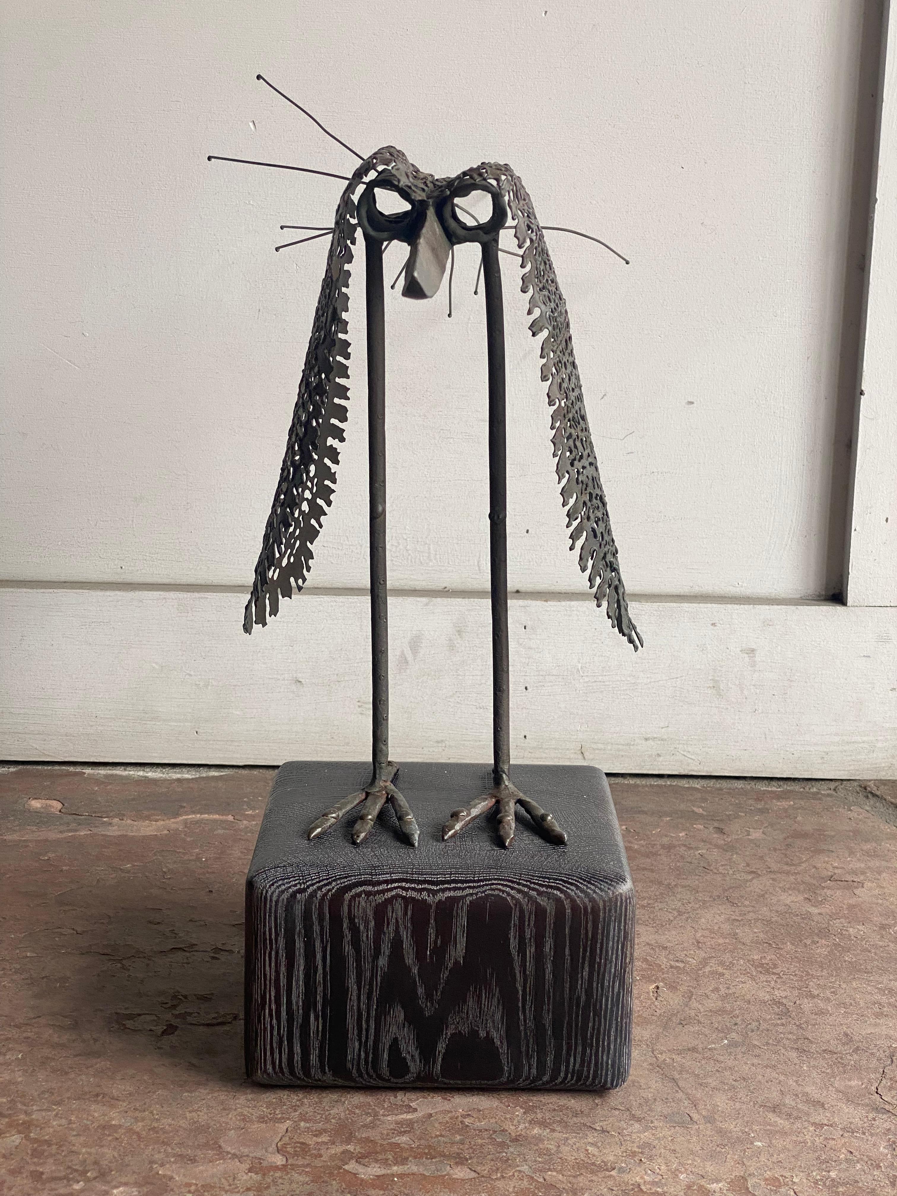 This metal bird sculpture features a stylized form with elongated legs and feet mounted on a dark-stained cerused base. The body is crafted from perforated metal sheets, giving a sense of lightness and texture, while the tail is composed of extended