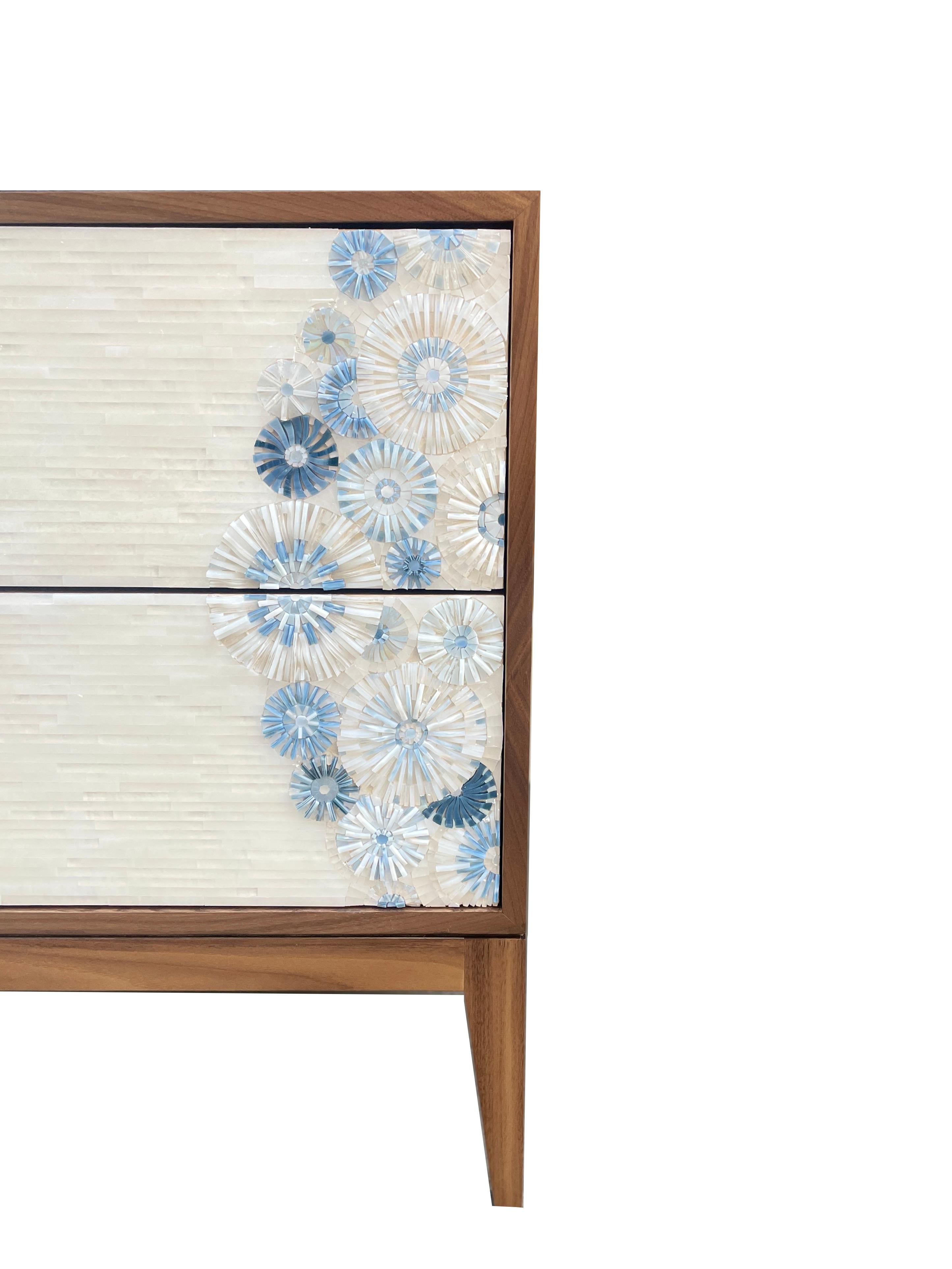The Milano nightstand by Ercole Home has a 2-drawer front, with Walnut wood in a Natural Finish. Hand cut glass mosaics in Blossom Pattern in a mix of blue colors decorate the surface along with a horizontal field of white glass stripe pattern.