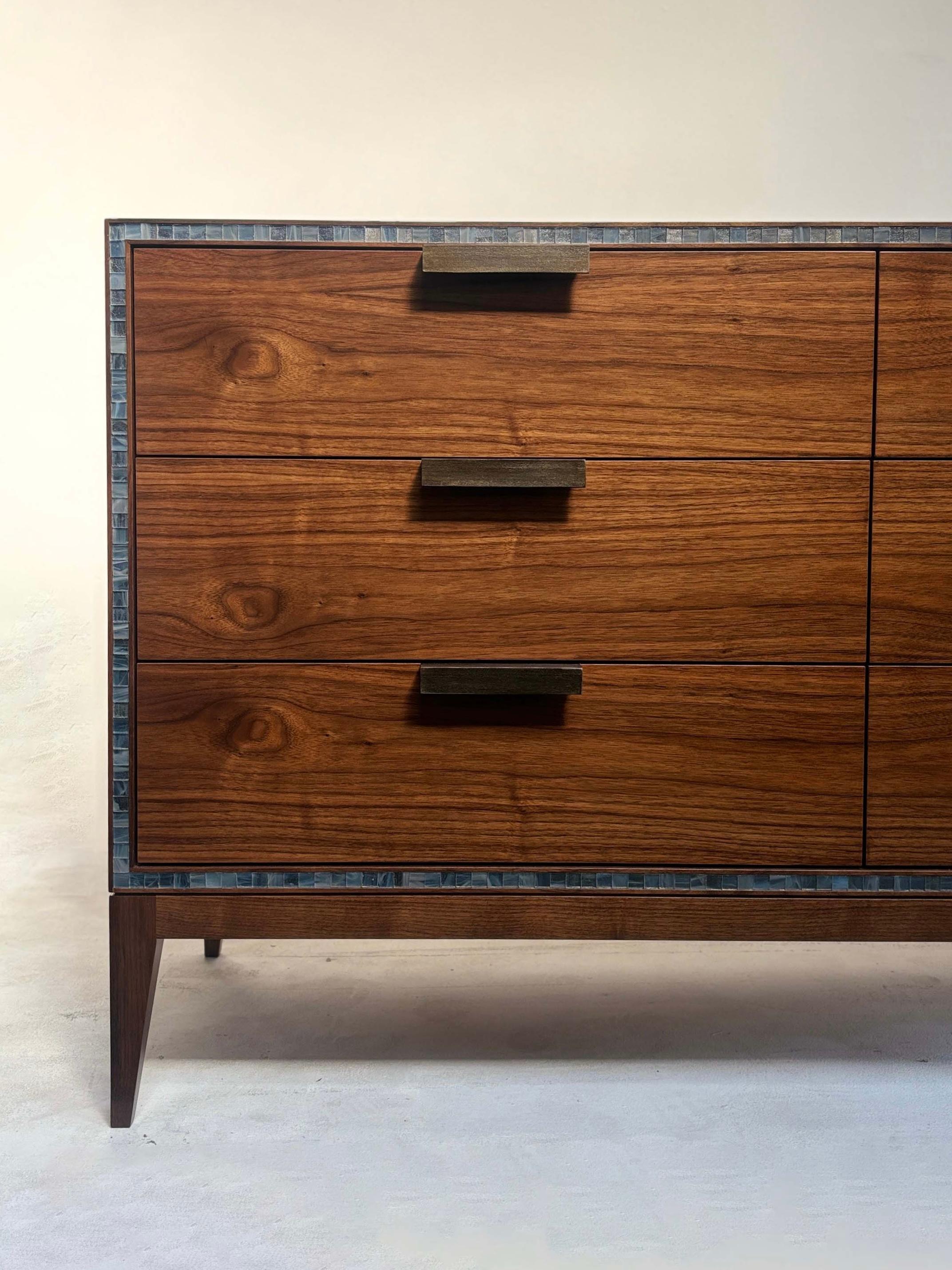 La cassettiera Milano di Ercole Home è composta da 9 cassetti tutti impreziositi da tiranti in acciaio industriale fatti a mano. La cassapanca è in noce nero americano con finitura Nature. Il bordo della cassapanca è delineato da un mosaico di vetro