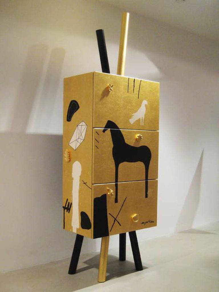 The limited edition cabinet designed by Mimmo Paladino is entirly covered in a layer of gold leaf and then decorated by hand with various designs in black and white by the author. The piece has three openings and offers ample storage. A mirrored