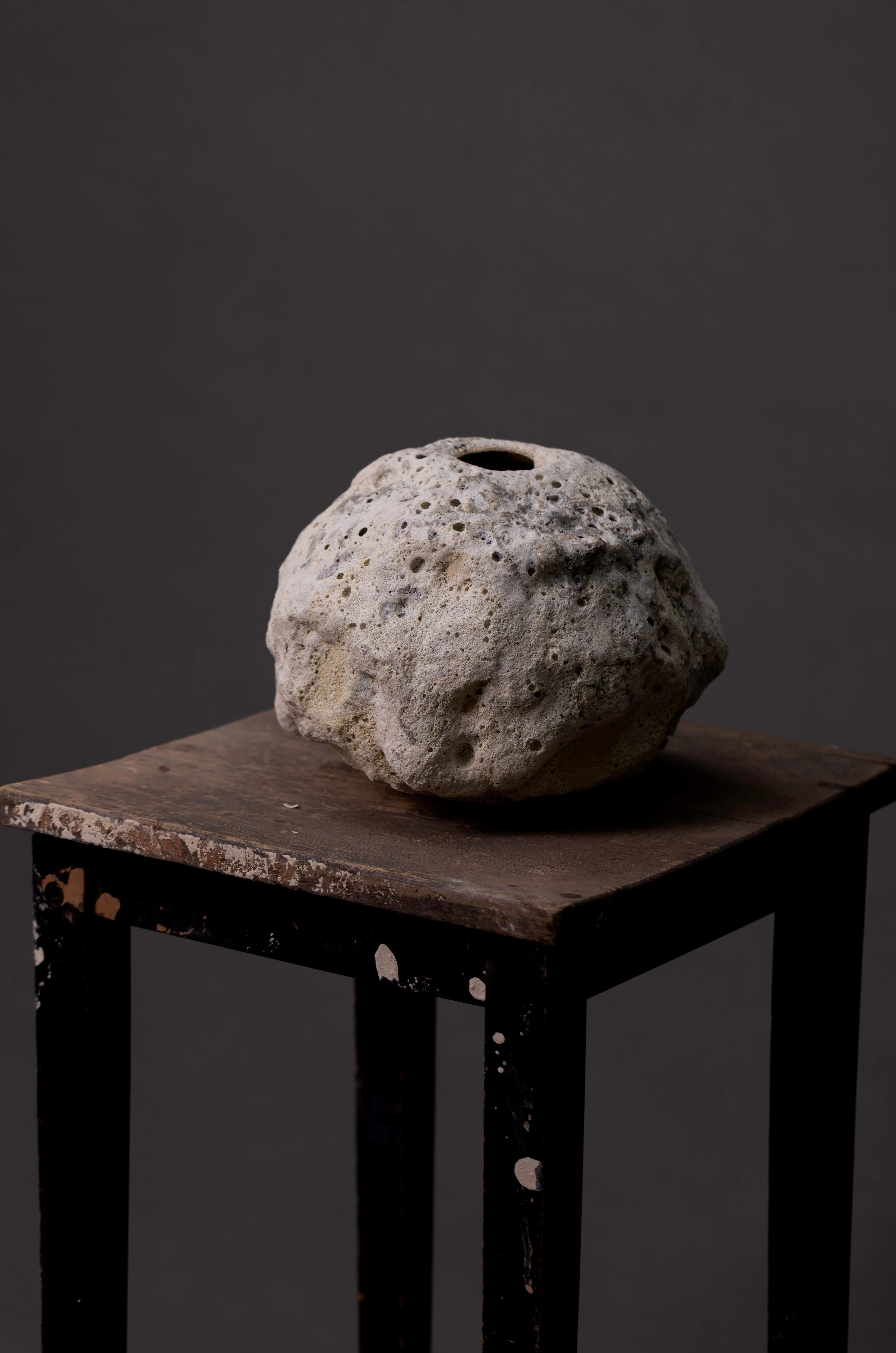 This handmade ceramic vase features a light beige finish and a roughly spherical form with a distinctly textured surface. Irregular pits and bumps create a porous, organic look, highlighted by soft variations in light and shadow. The matte glaze