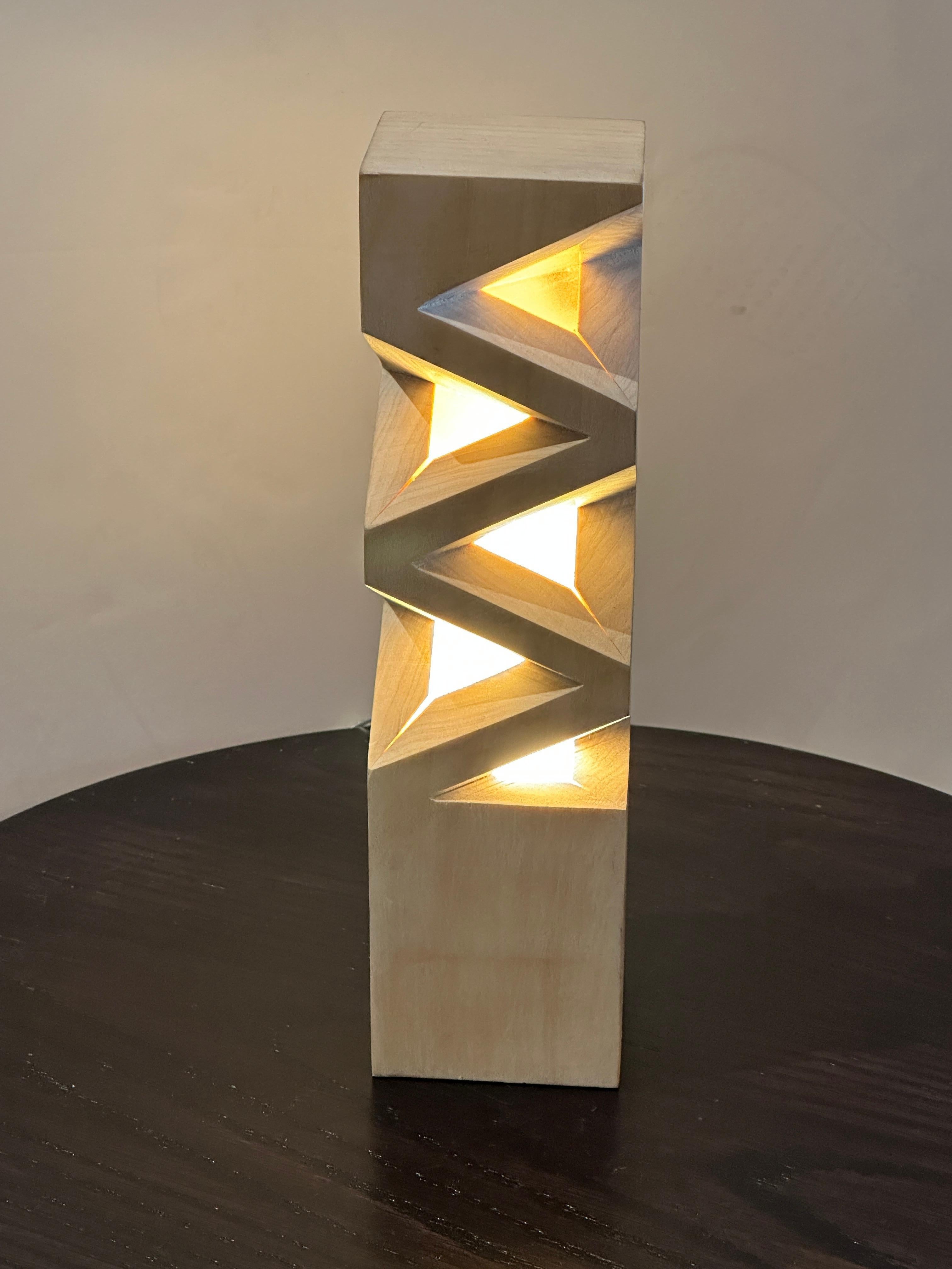 This modern Minimalist geometric natural wood table lamp features three tetrahedron shapes carved on two sides and two tetrahedron shapes on the other two sides. Crafted from light-colored wood filled with resin to spread the light, the geometric