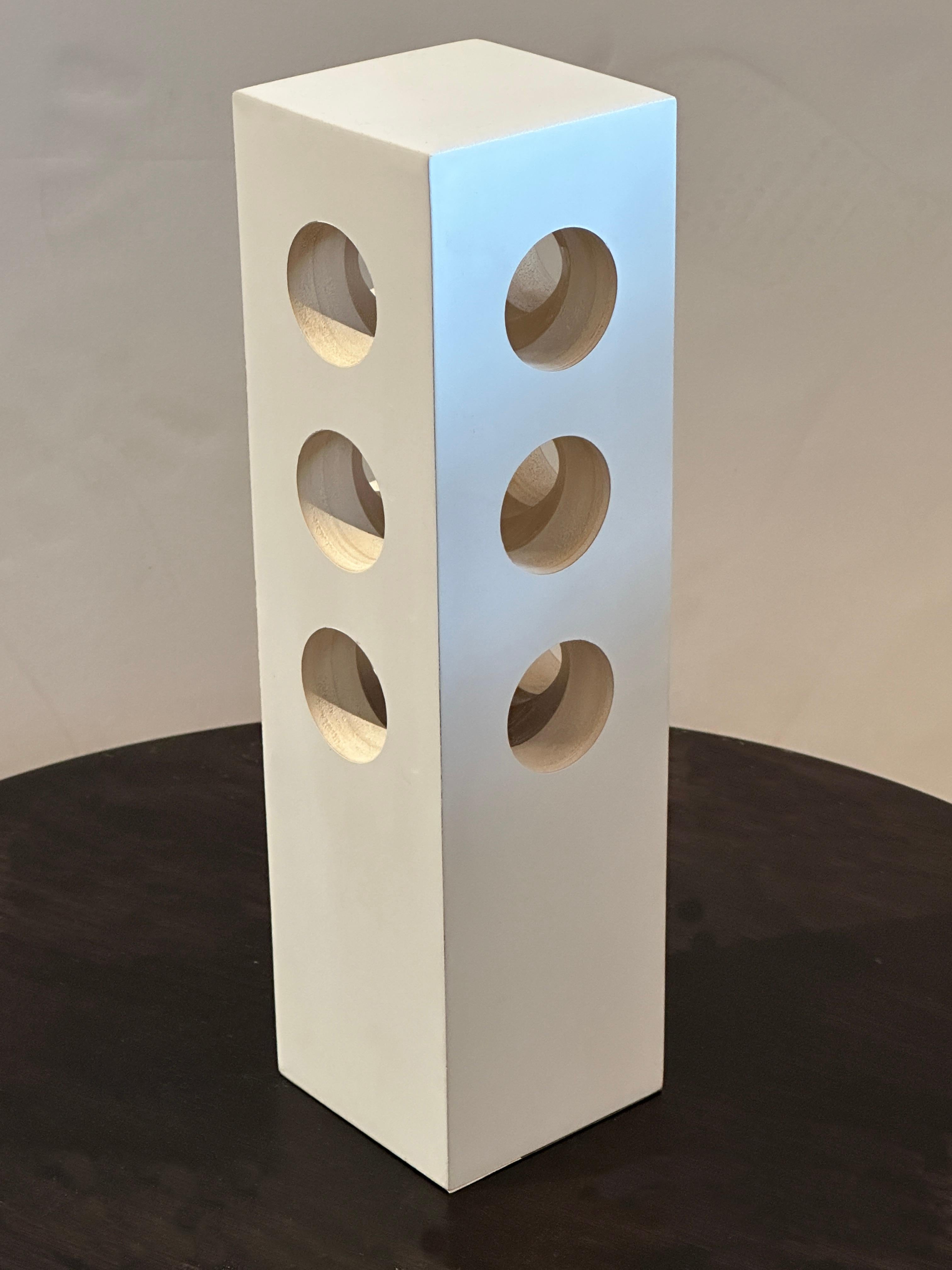 This modern Minimalist white table lamp features three regular holes on each side, allowing soft and calming light to pass through. The lacquered white wood finish ensures that it blends seamlessly with any color scheme. This table lamp will