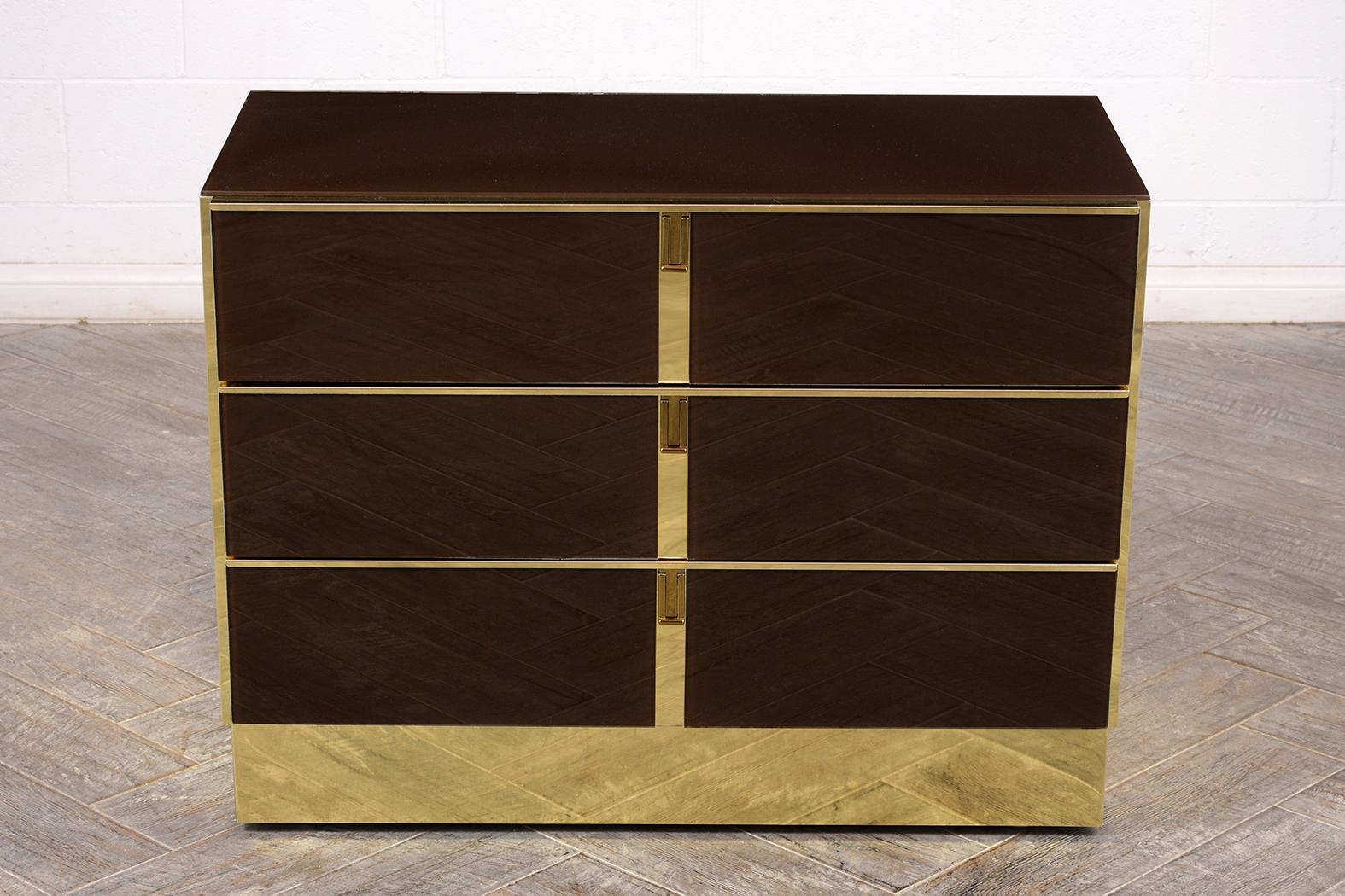 This 1970s modern brass and smoked glass chest of drawers features a glass top and three large drawers. Each drawer has two glass panels and brass pulls at the center. This chest of drawers rests on a floating base. The chest is sturdy and ready to