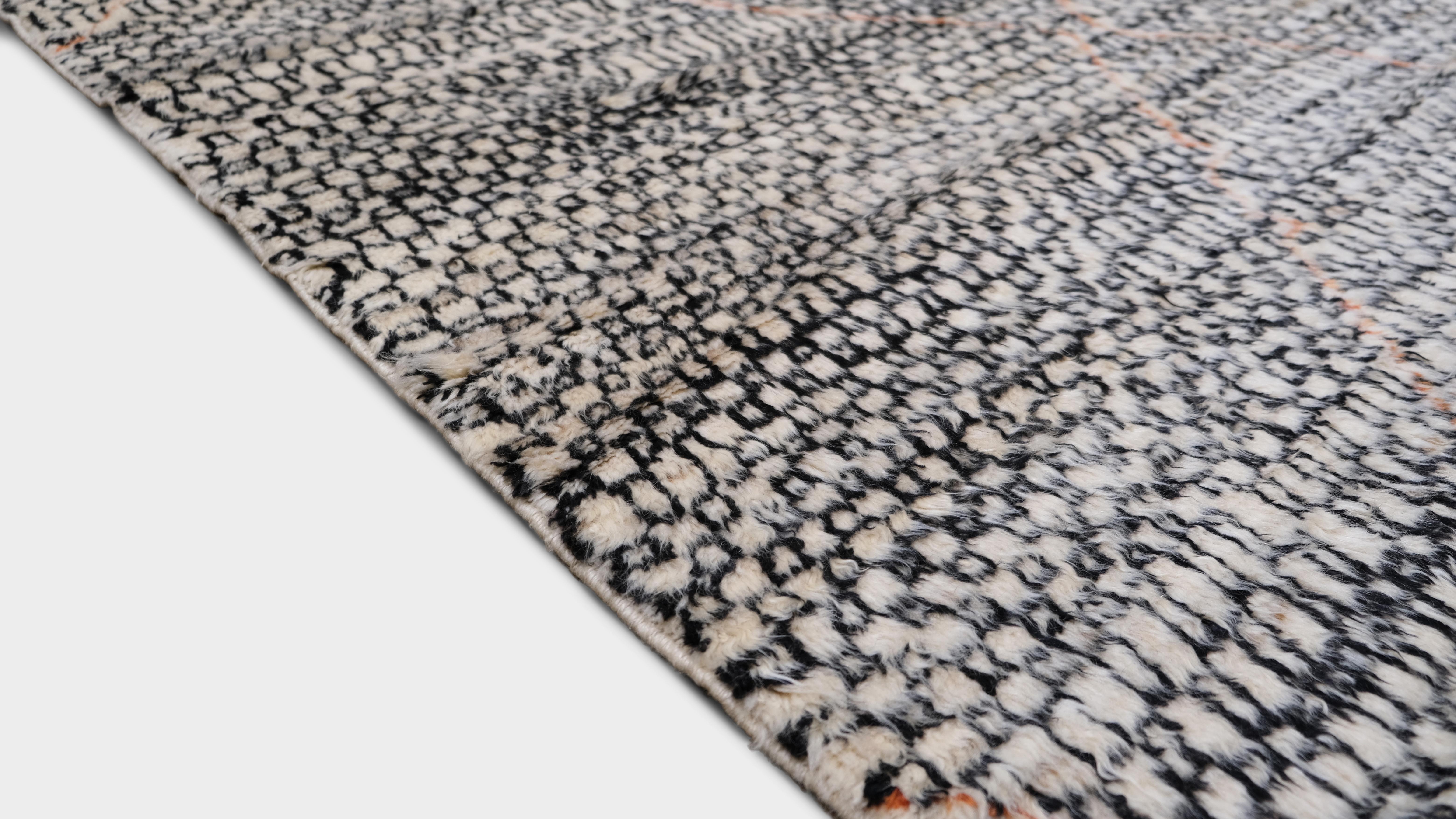 Dieser handgeknüpfte Wollteppich ist eine raffinierte Verschmelzung von modernem Design und Berbereinflüssen. Er zeigt ein dynamisches Rautengittermuster in Schwarz und Elfenbein mit dezenten Korallenakzenten. Seine einzigartige strukturierte