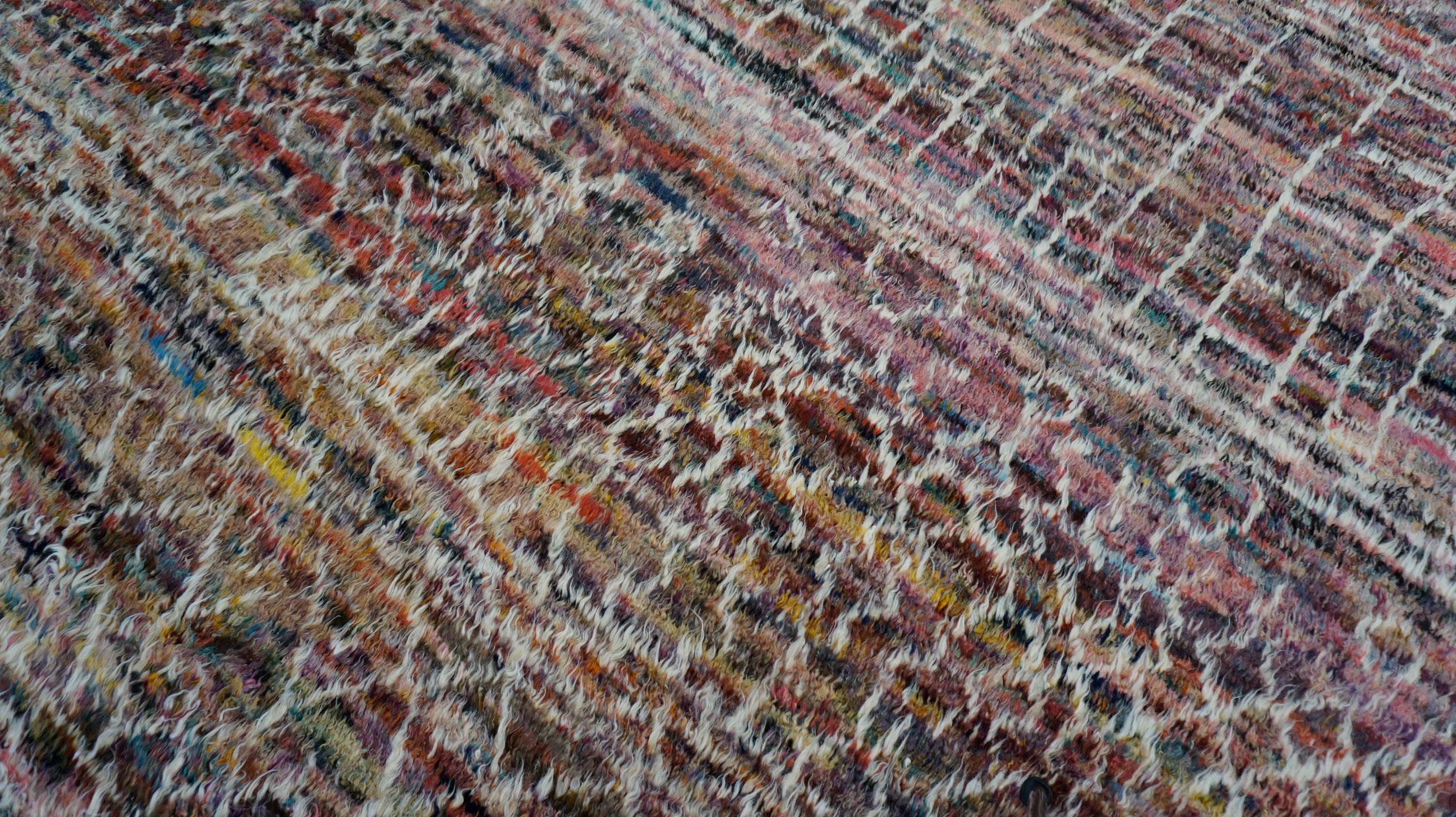 This stunning hand-knotted wool rug showcases a vibrant mix of earthy tones layered with intricate tribal-inspired linear motifs. The design features a richly textured surface, created with a dynamic interplay of pink, purple, brown, and white
