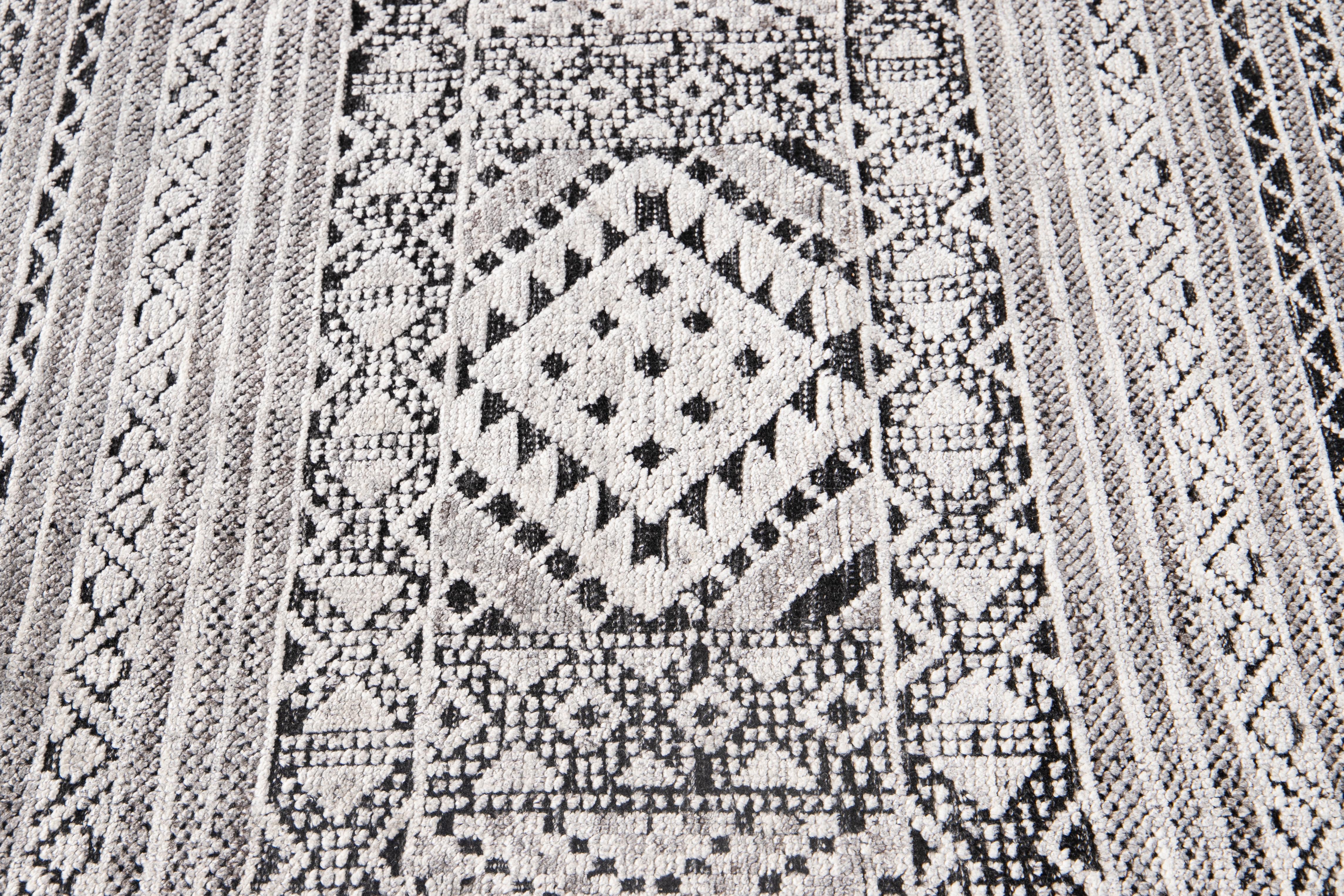 This Indian rug boasts a stunning texture and high-low pile, handcrafted from premium wool with tasteful Ivory highlights. The light gray and black background showcases an intricate, all-over geometric motif that will elevate any room's