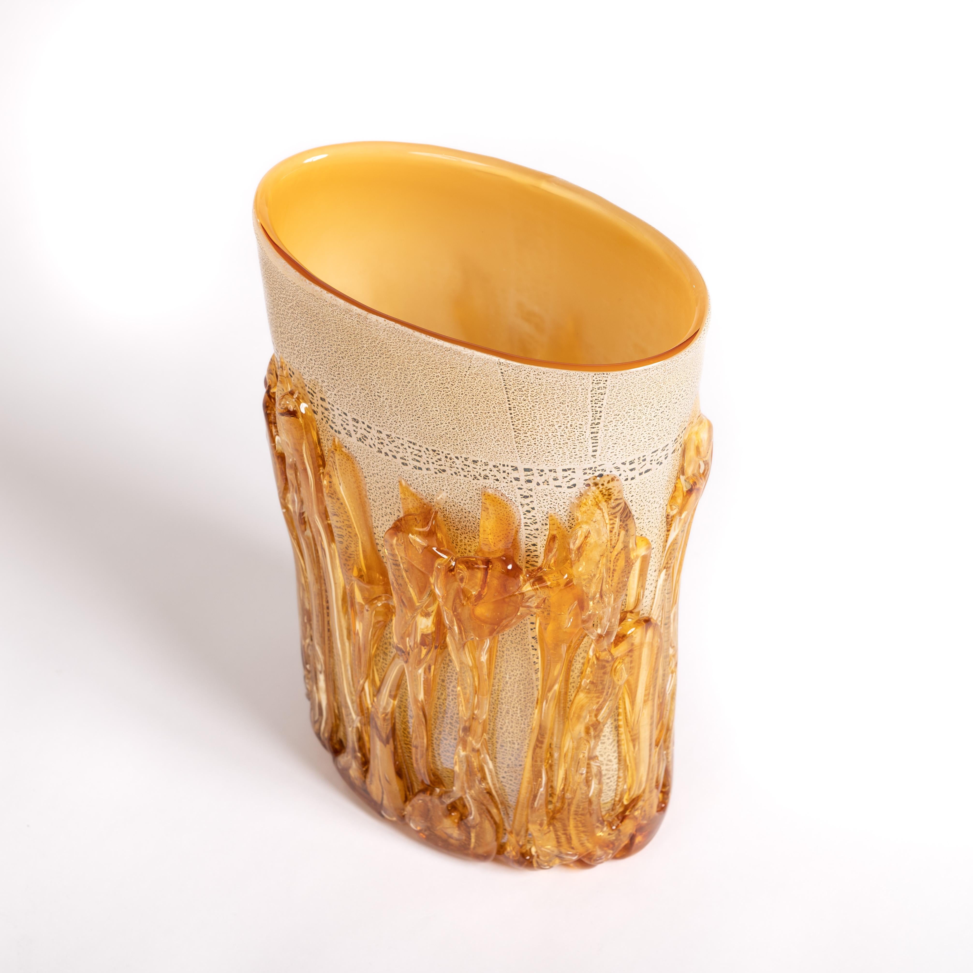 A straight-lined vase with a matt, amber-colored interior and an exterior covered with an abstract and porous-looking gold glaze.
Strands of glass are laid all around it, these strands are slightly thicker at the top and bottom and also have a
