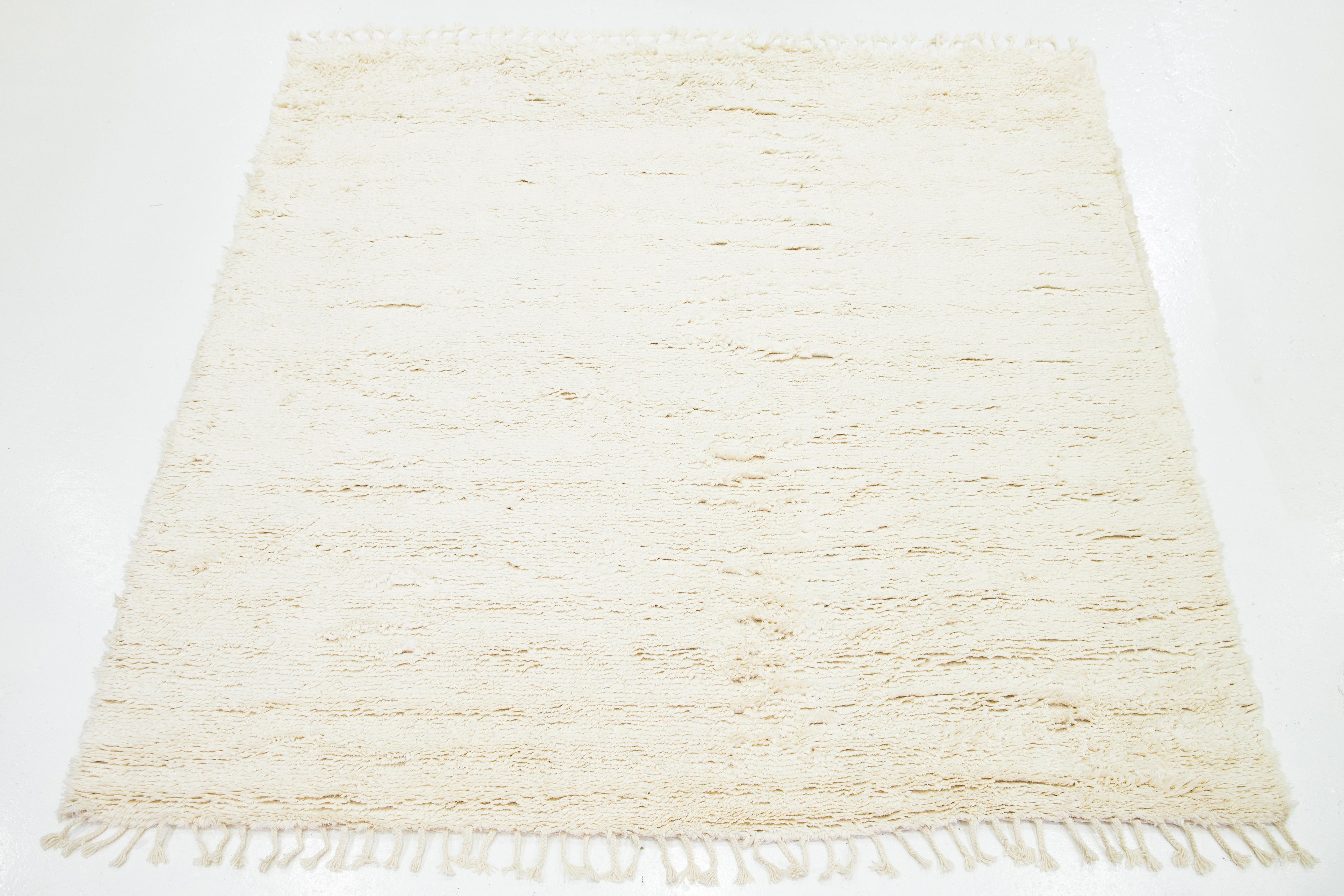 Dieser moderne, handgeknüpfte Wollteppich im marokkanischen Stil zeichnet sich durch ein elfenbeinfarbenes Feld und Fransen mit einem plüschigen, soliden Design aus.

Dieser Teppich misst 8'6
