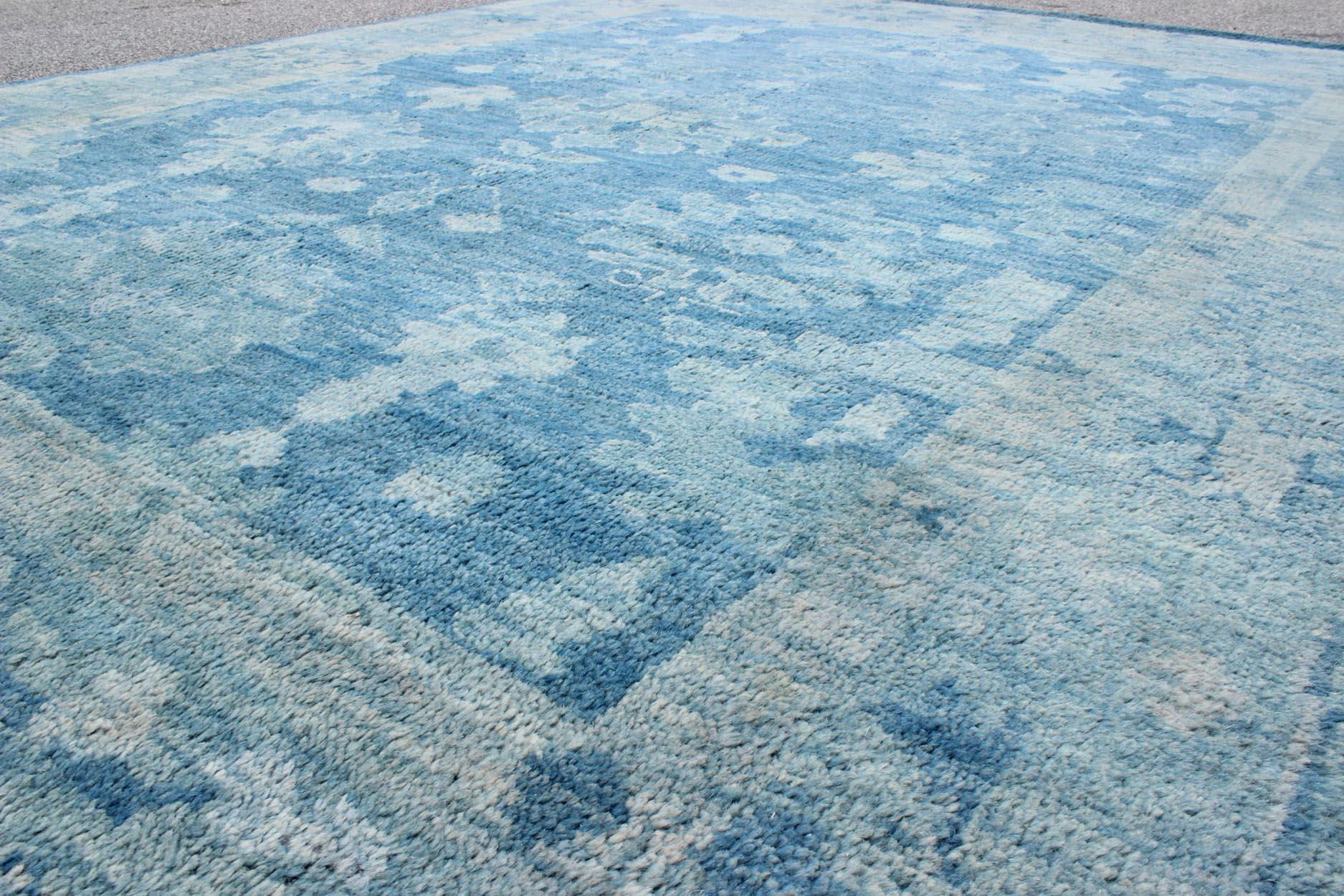 This modern Indian Oushak rug, made by Keivan Woven Arts, has been hand-knotted in wool and features an all-over, sub-geometric floral design rendered in blue tones. A complementary, multi-tiered border encompasses the entirety of the piece; making