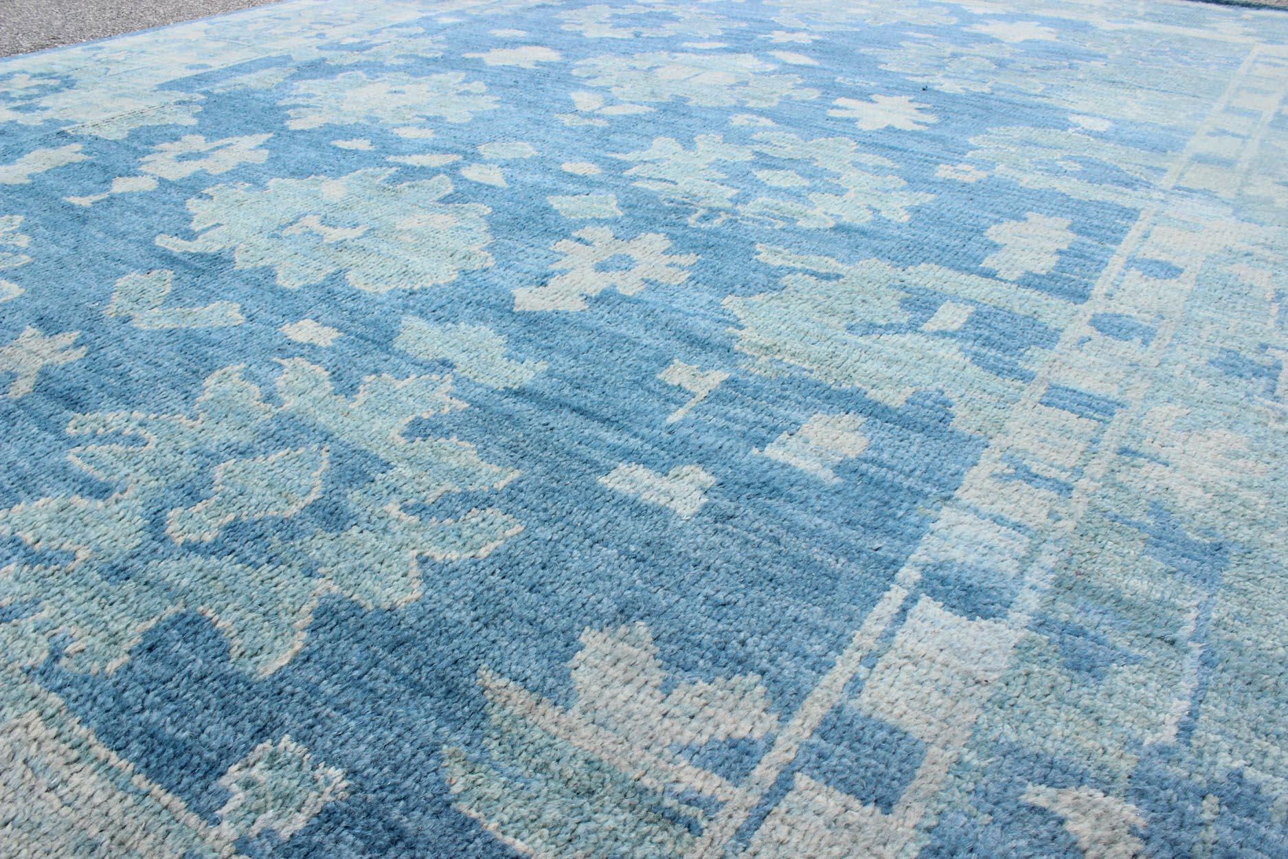 Questo moderno tappeto indiano Oushak è stato annodato a mano in lana e presenta un disegno floreale sub-geometrico all-over nei toni del blu. Un bordo complementare e a più livelli abbraccia l'intero pezzo, rendendolo adatto a un'ampia varietà di