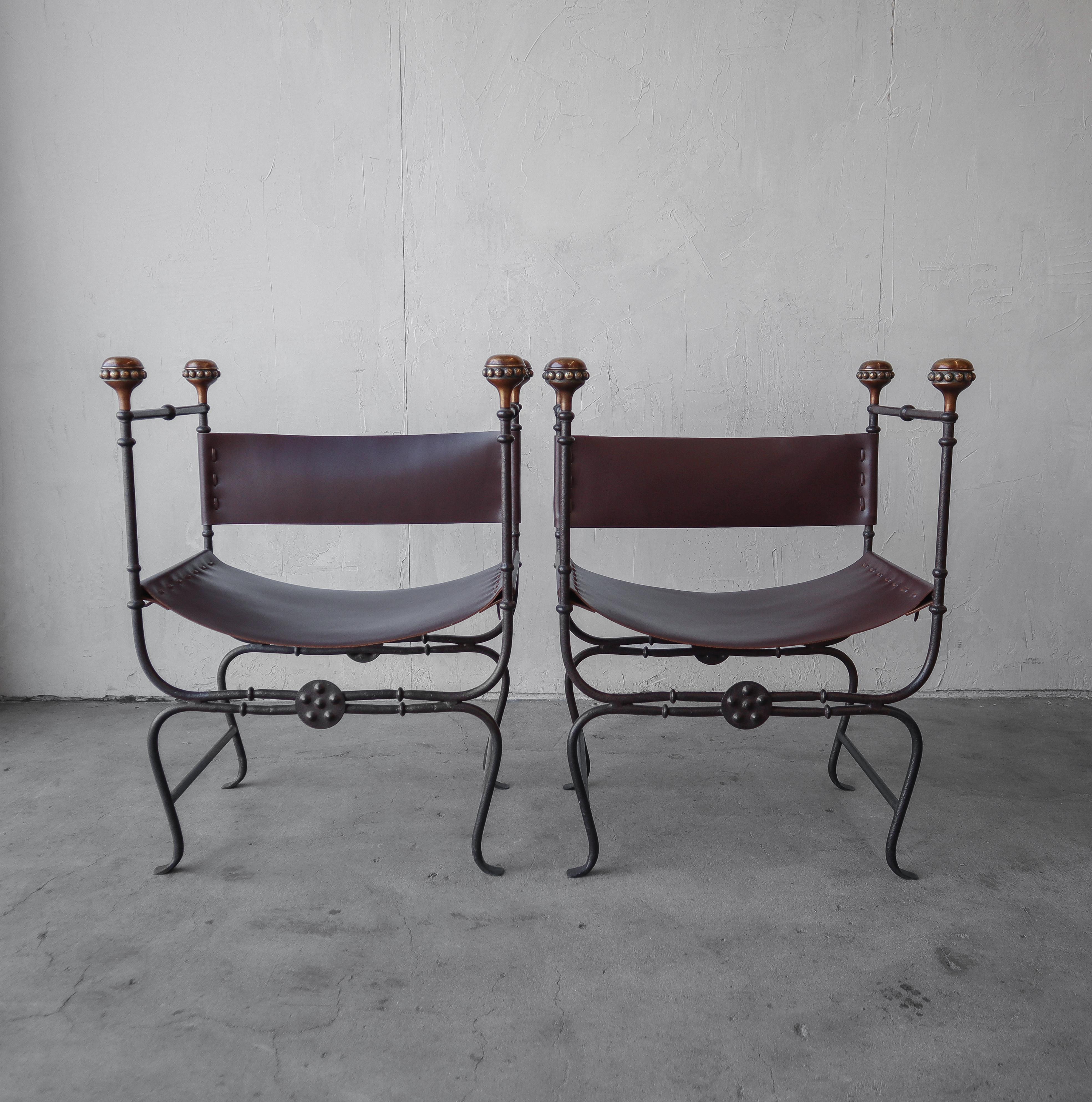 Great modern pair of iron, brass and saddle leather curule savonarola chairs. These chairs are heavy iron construction with nice patinaed brass finial details and thick leather sling seats and backs.

Chairs are in excellent condition with no
