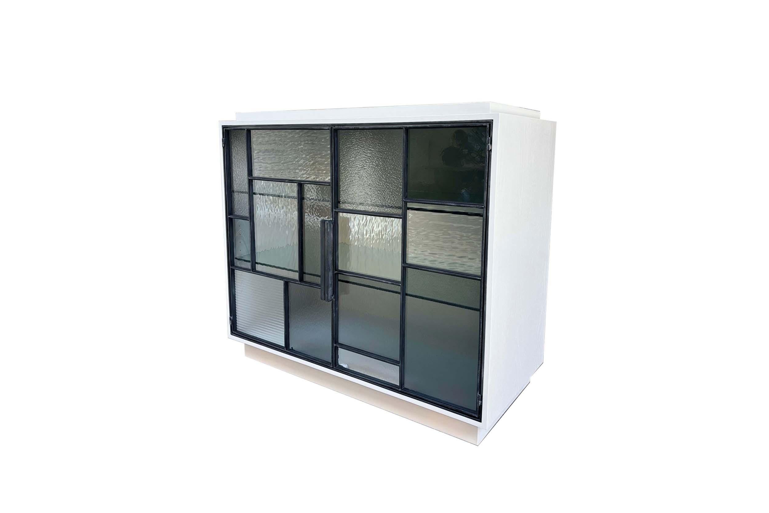 The Modern Platform Dry Bar Cabinet features two doors outlined in blackened metal that frames a unique combination of hand cut glass all varying in size, color, and texture. The exterior of is finished in Misty Grey on Oak and the interior is
