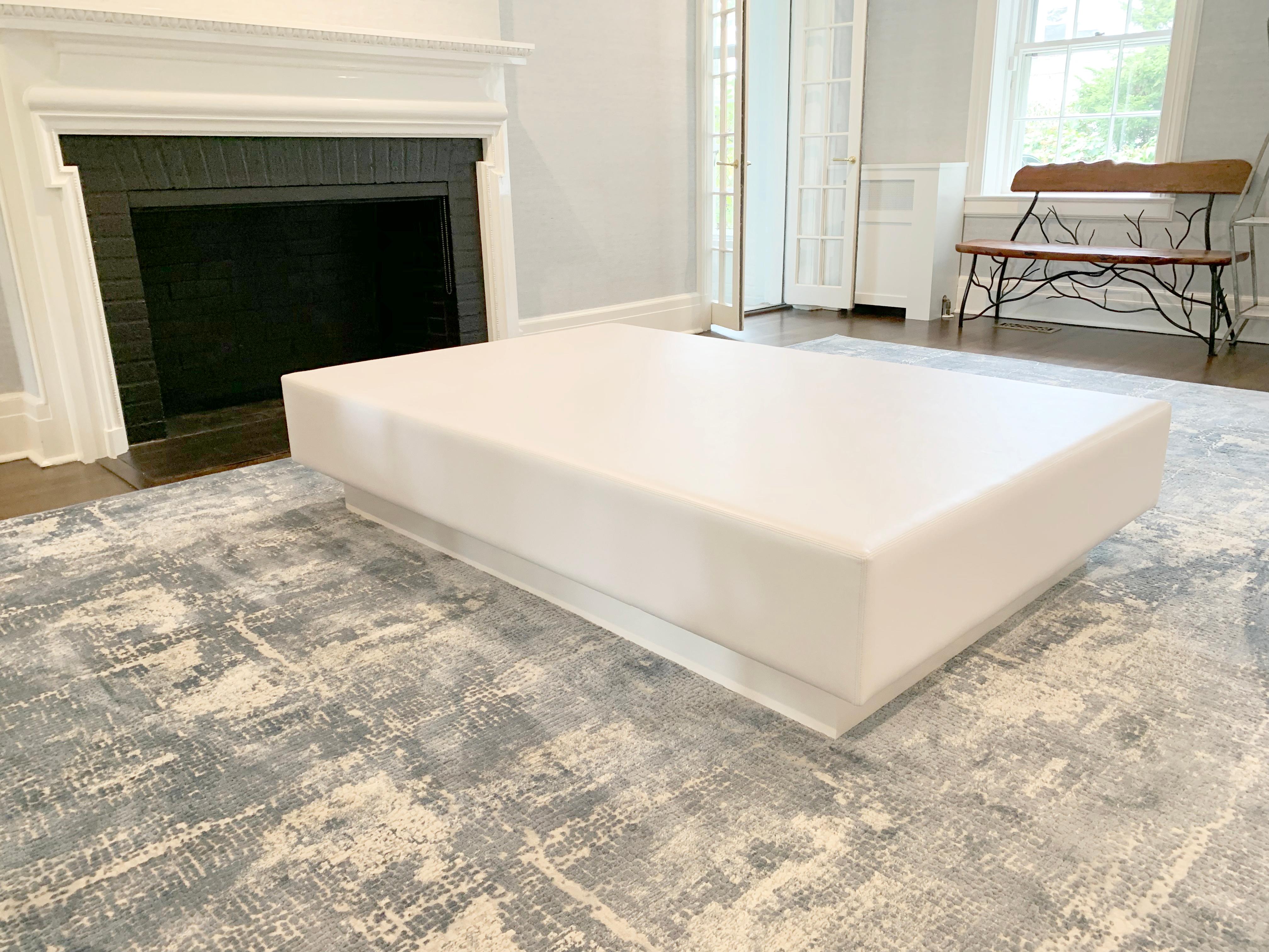 A long rectangular coffee table inspired by simple and modern design. The top is lightly padded and wrapped in a vinyl fabric to soften hard edges and protect against spills. The wooden base is lacquered in white dove and recessed for a floating top