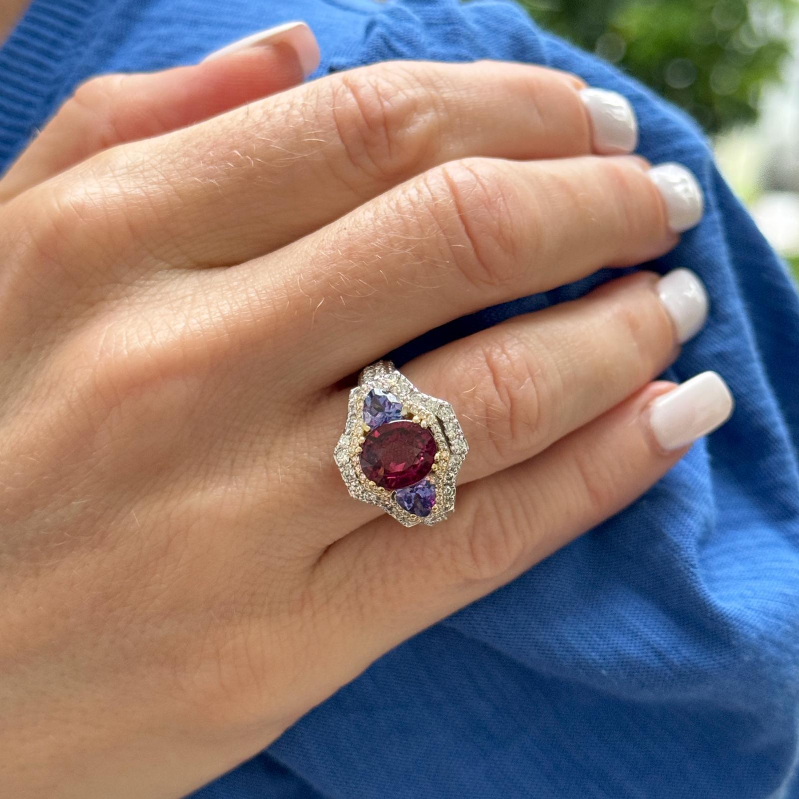 Questo anello da cocktail moderno presenta un sorprendente granato rodolite da 3 carati al centro, affiancato da due vibranti gemme di tanzanite (per un peso totale di 0,60 carati). I ricchi colori sono ulteriormente accentuati da un'aureola di