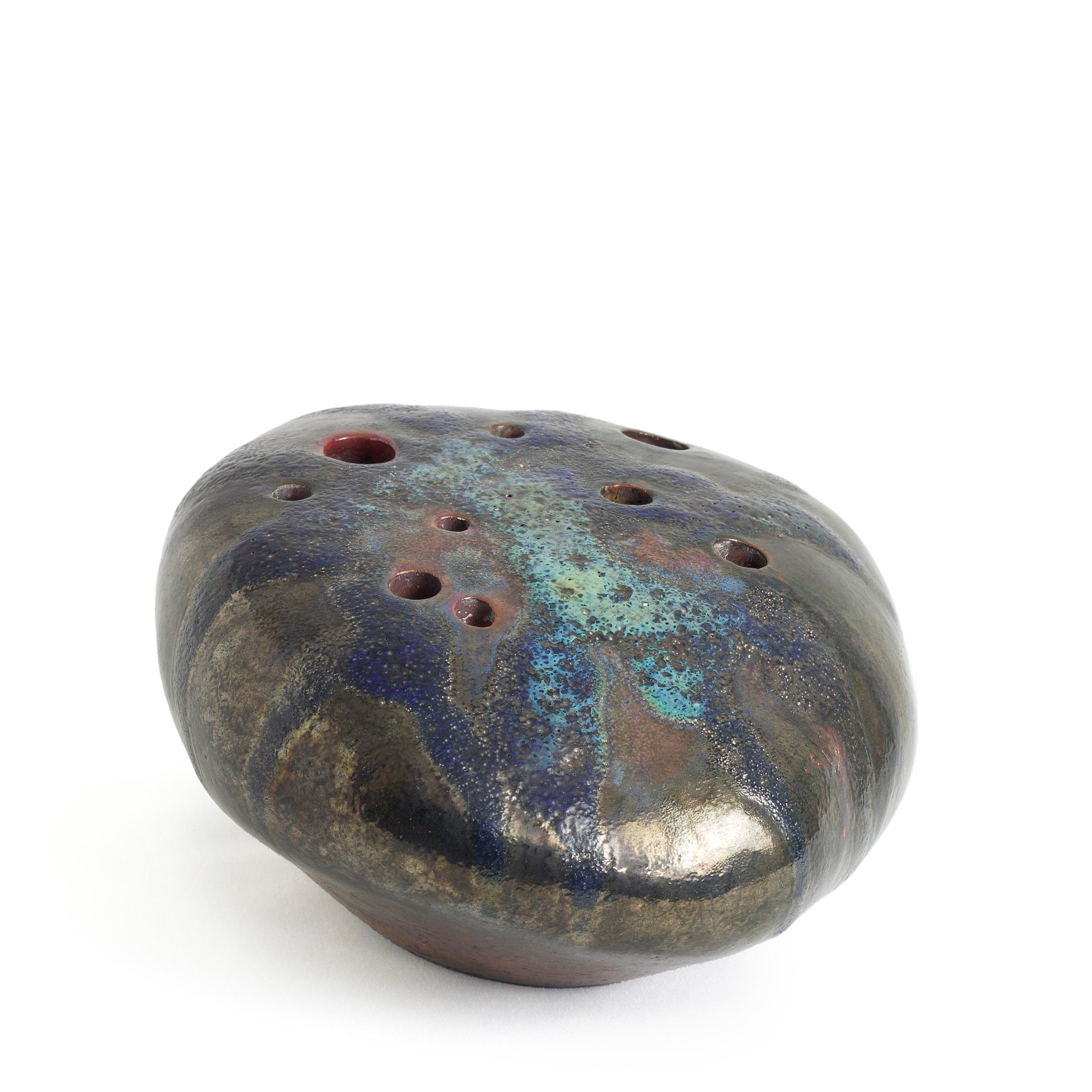 A Ceramic Raku fired rock shaped sculpture. Designed as an ikebana, and an incense holder, this ceramic peddle shows an interesting pattern of holes and green copper strains, highlighted by the dark metal predominant color that turns into oxide iron