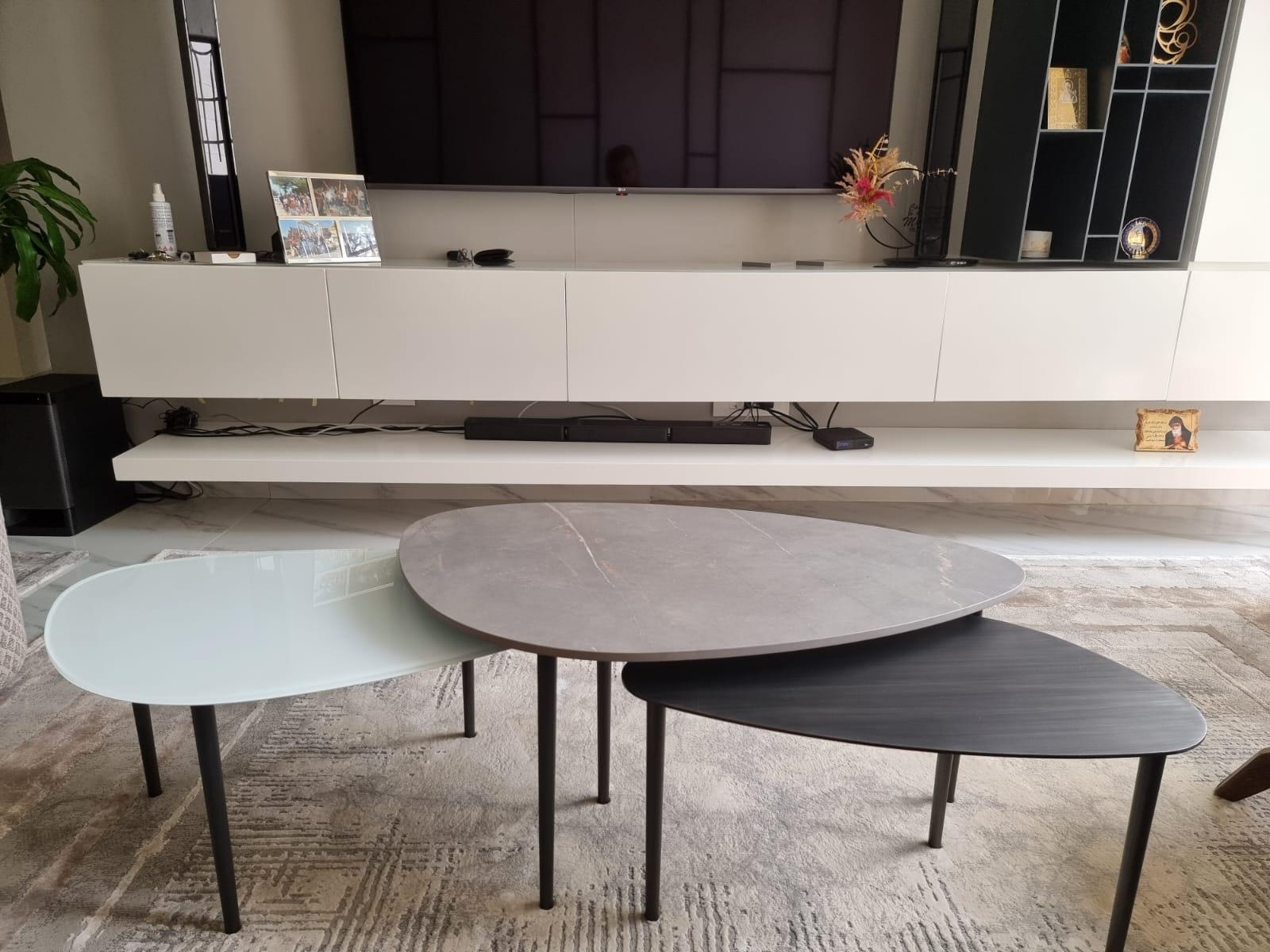 Set of 3 coffee tables that have the same top shapes and legs but in different sizes, varied heights and several materials of the tops. The 3 tops vary between back-painted glass, grey ceramic and brushed gun metal. 
The tops have asymmetrical oval