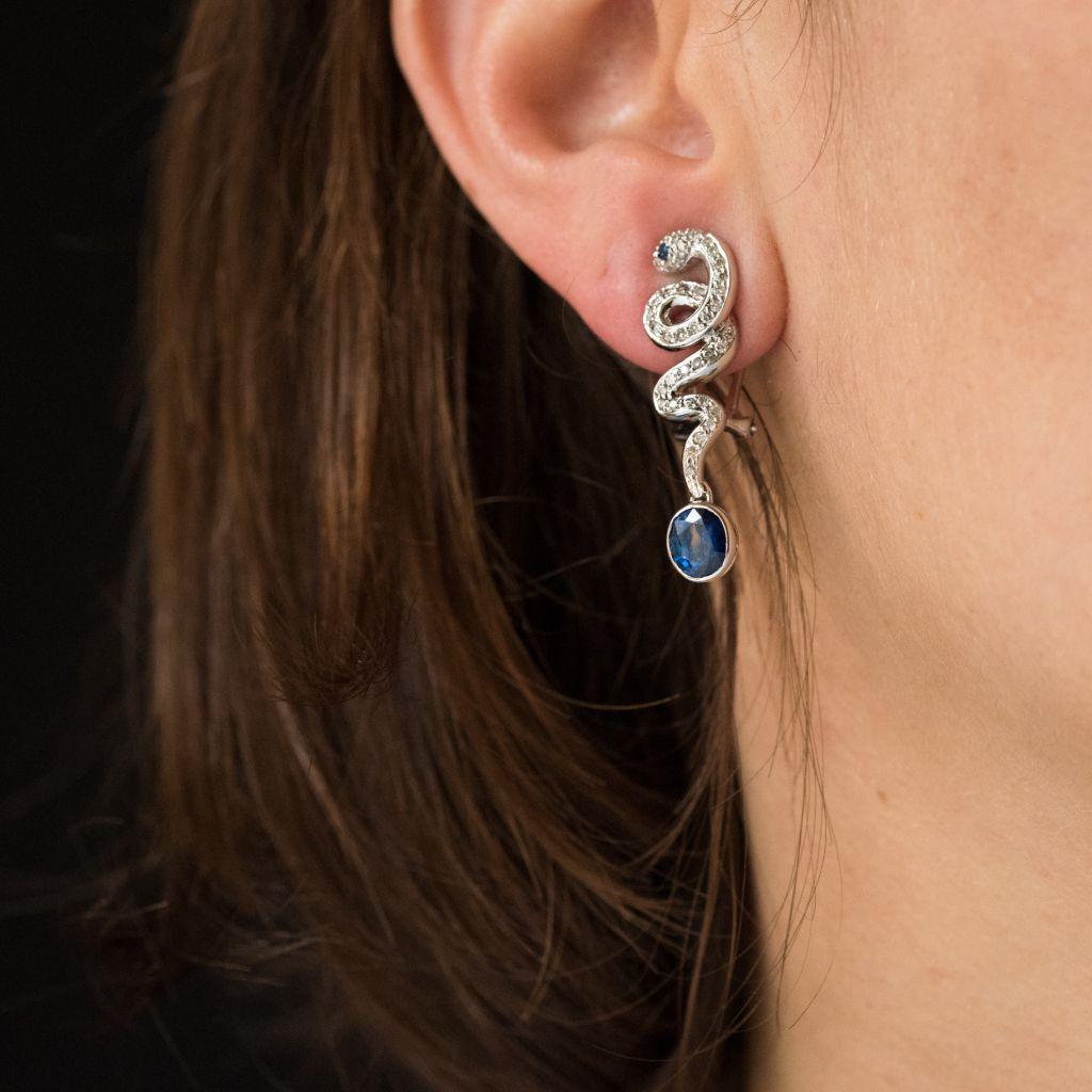 For pierced ears. 
Pair of 18 carat white gold pendant earrings.
They feature curled snakes, each with diamond pave and a bezel set sapphire at the base. The eyes are small sapphires. The earring clasps feature safety hooks. 
Total weight of