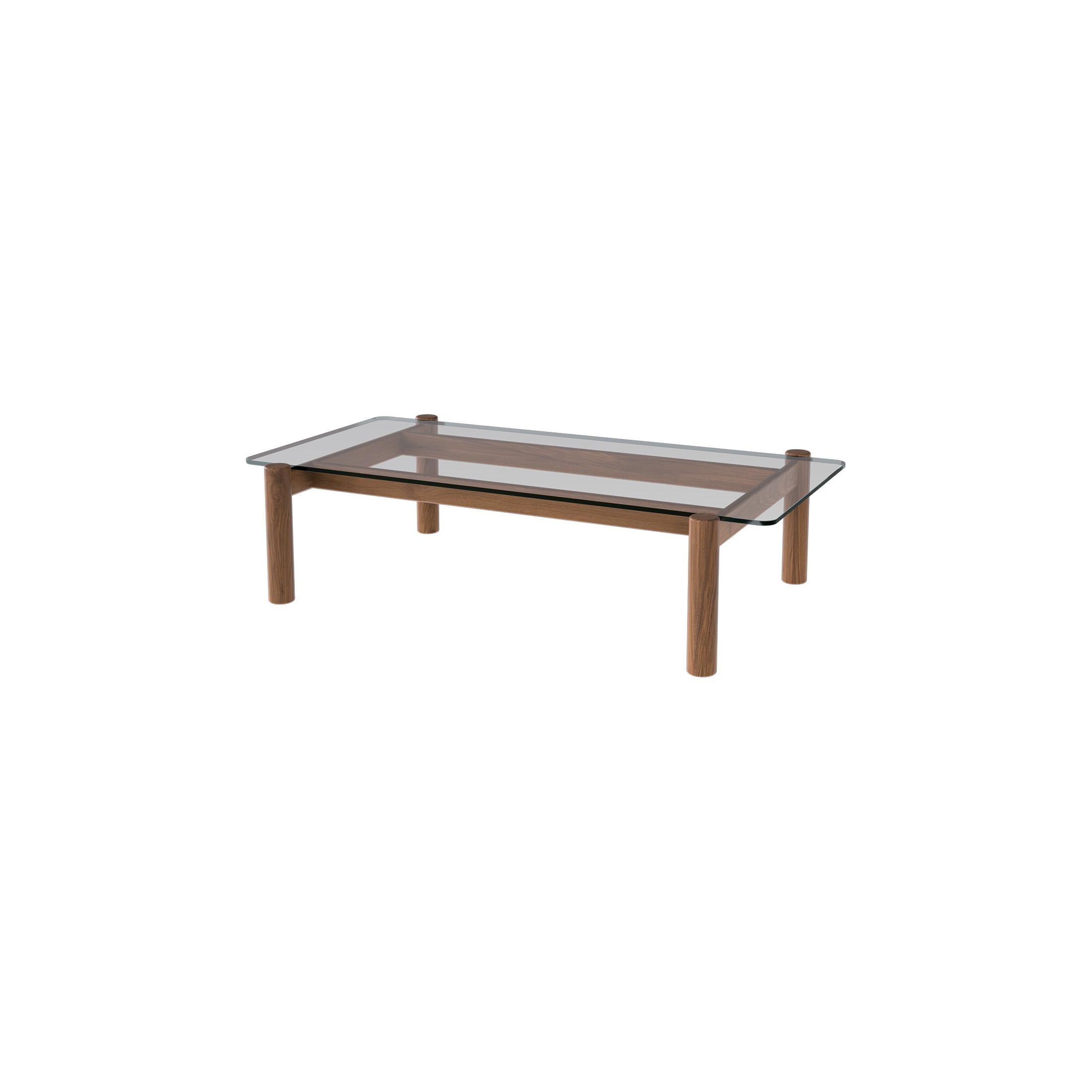 This elegant coffee table features a clear plate glass top elegantly framed by a distinctive asymmetric pinwheel base. Crafted entirely from solid Ash or Walnut, it showcases oversized, hand-turned legs with refined exposed through-tenon joinery.