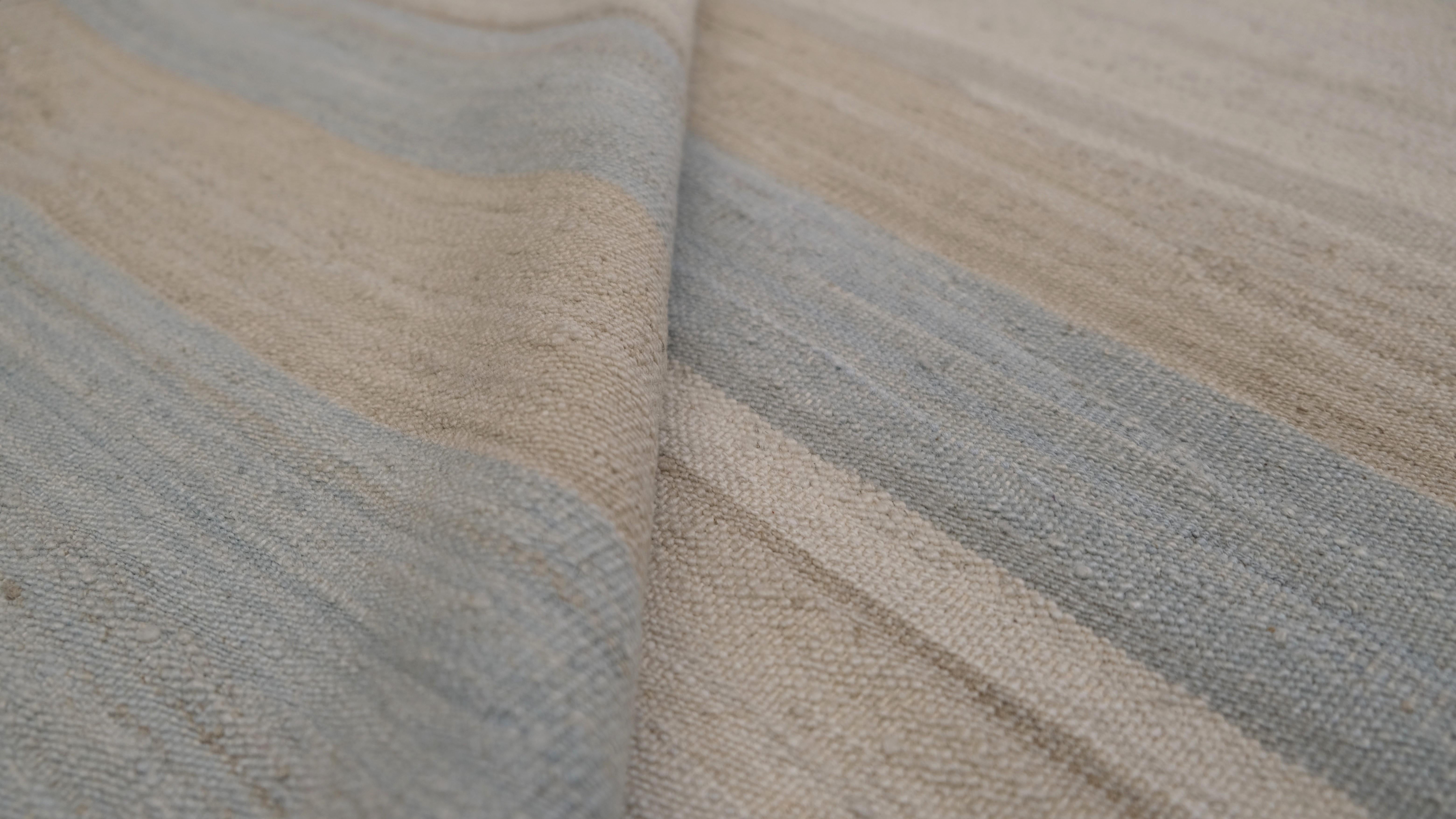 Ajoutez une touche d'élégance sereine à votre espace avec ce kilim exquis, doté d'un motif dégradé rayé moderne. Le jeu subtil de bandes horizontales en beige doux, ivoire et bleu sourd crée une esthétique contemporaine apaisante qui rappelle les