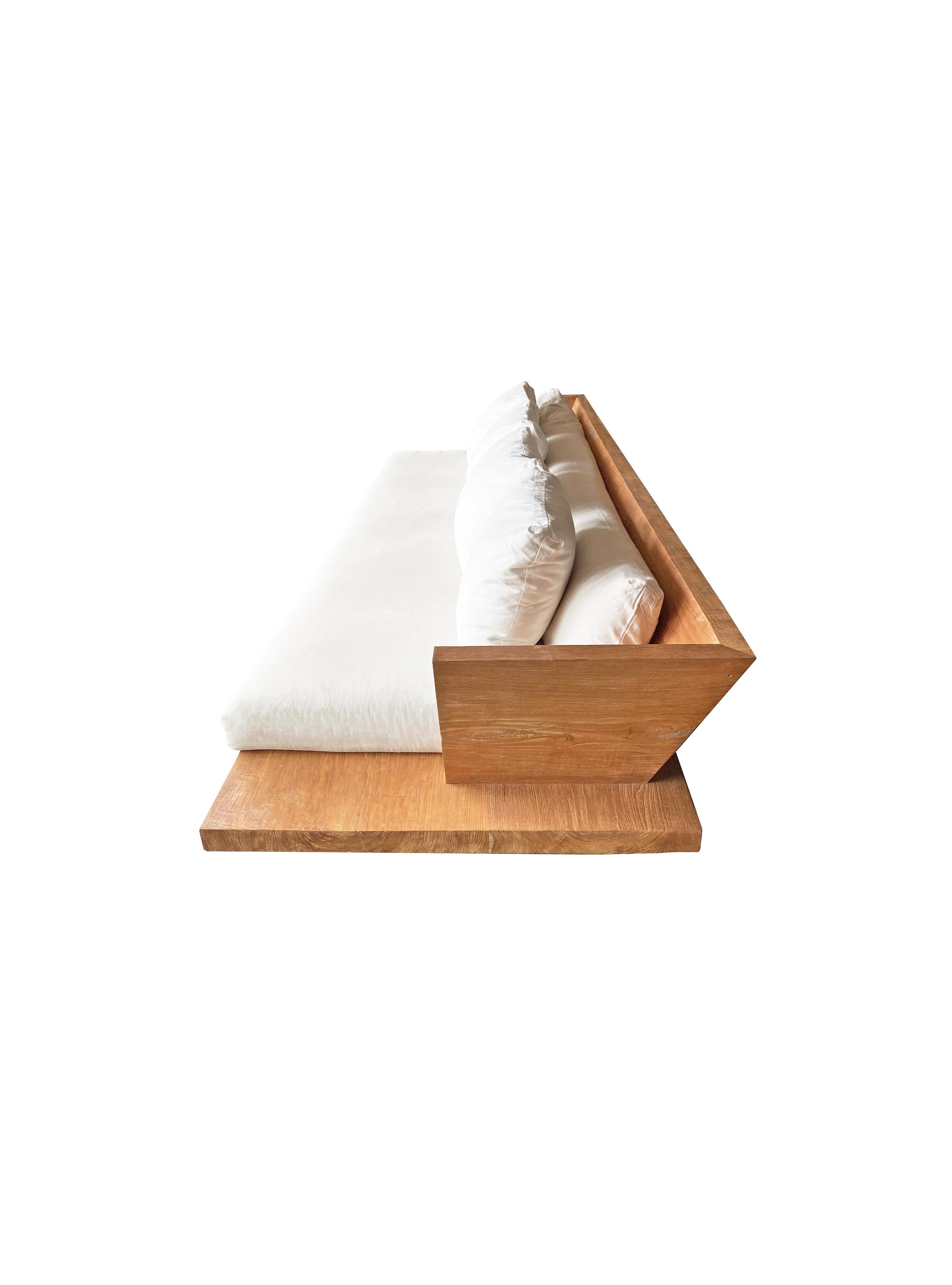Ce canapé moderne en bois de teck présente une grande base en dalles de bois de teck, un dossier et est surélevé par deux blocs de bois de teck peu profonds. Le canapé est livré avec des coussins qui s'étendent sur toute la longueur du dossier et de