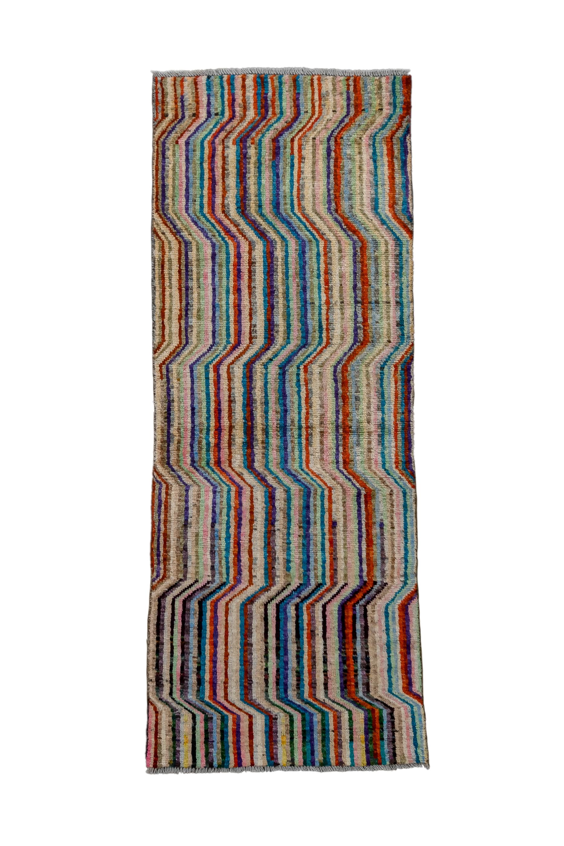 This long pile, elongated mat, shows a pattern of conforming, compressed, colourful trapezoidal meanders. No field tone, but a palette including:  luminous light blue,; sky blue; red; rust; beige; and ecru. Eye-dazzler pattern runs end to end and