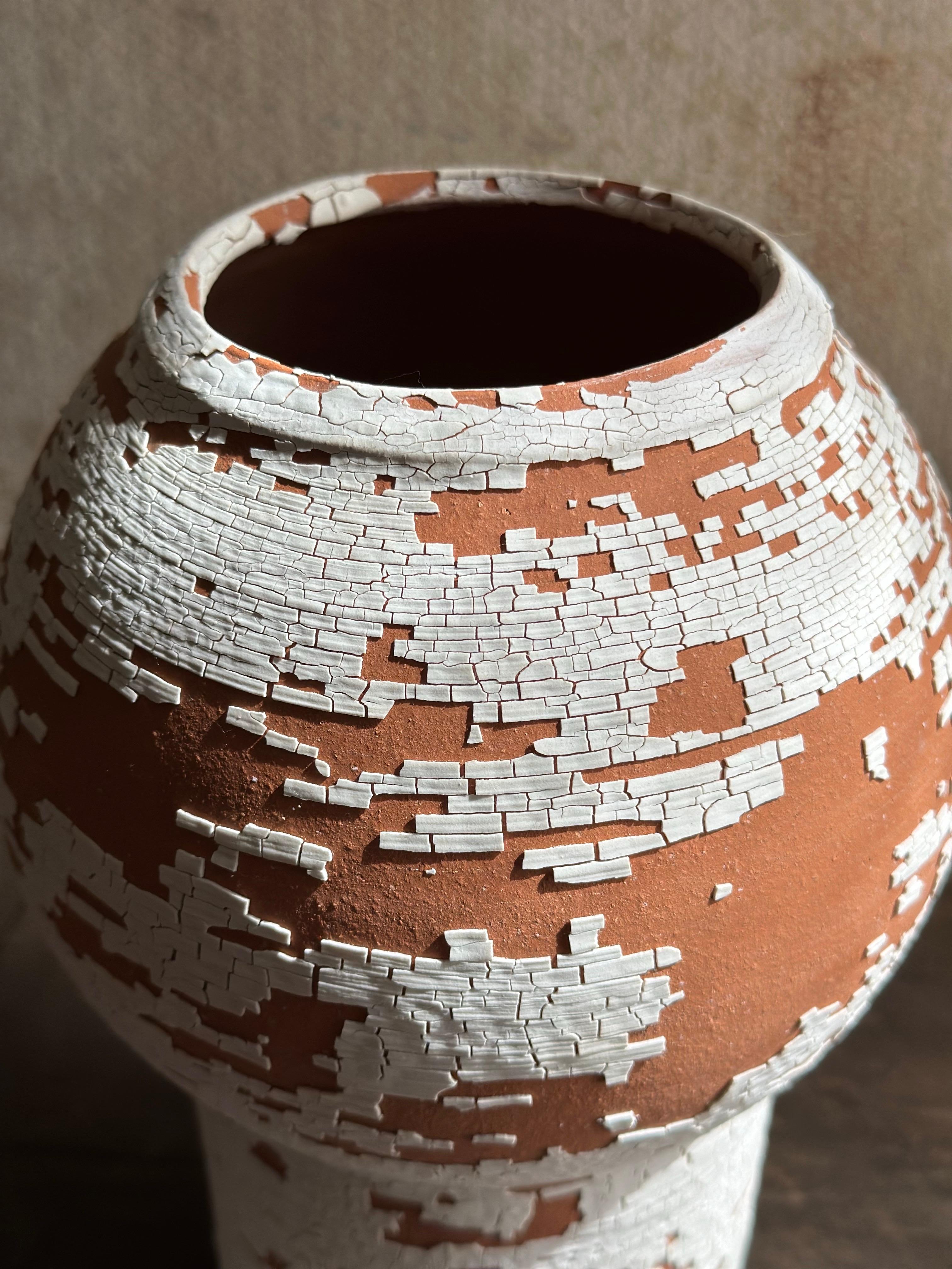 Vaso in ceramica realizzato a mano con una forma scultorea e audace e un aspetto grezzo e terroso. Lo smalto bianco crepato rivela strati di argilla rossa, creando una texture dinamica che sembra naturale e senza tempo. Questo vaso, perfetto per