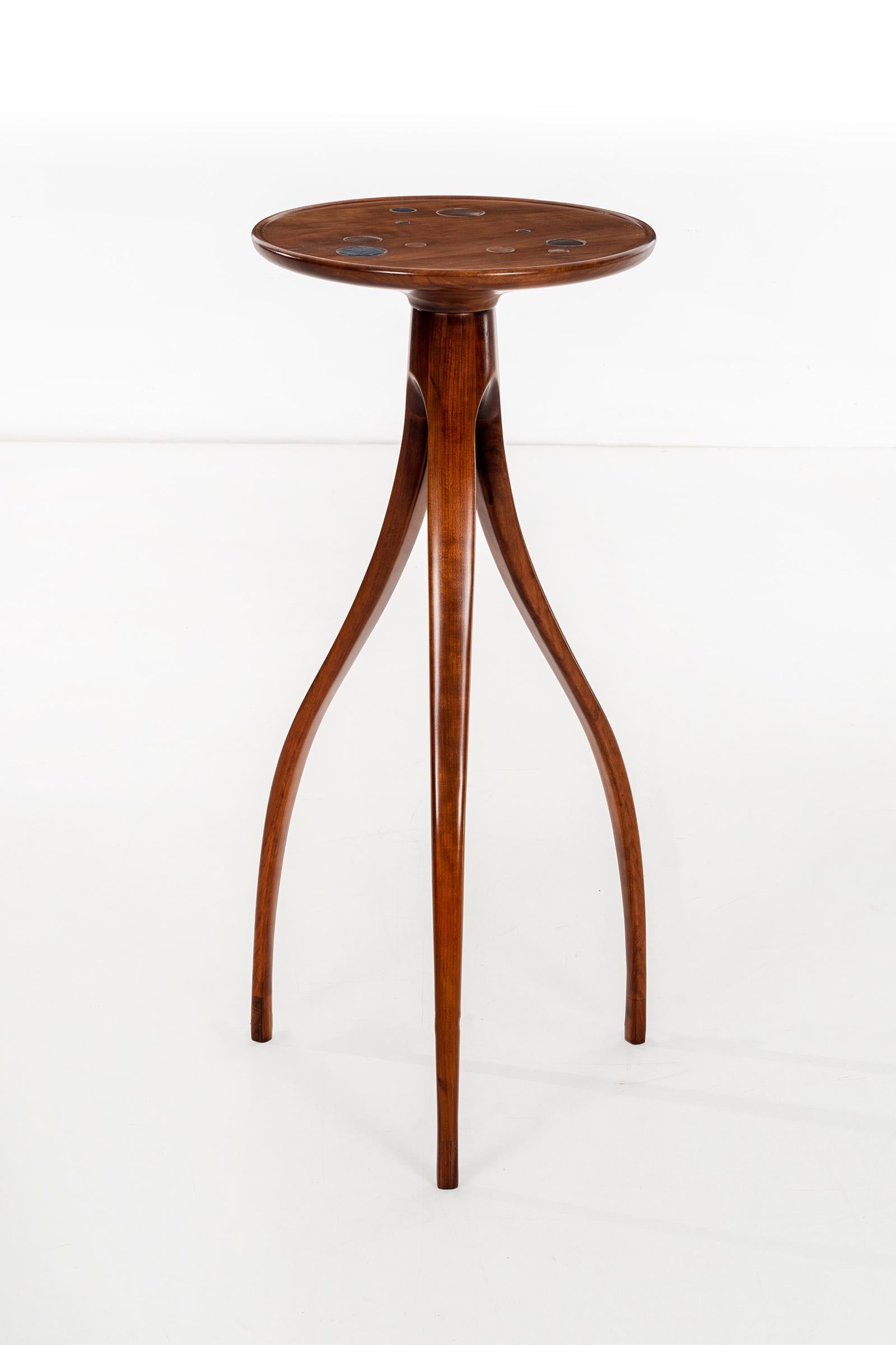Slender scale pedestal high tripod table, solid carved walnut lip edge top with ceramic design details inset.
Feet have lighter tone wood accent.