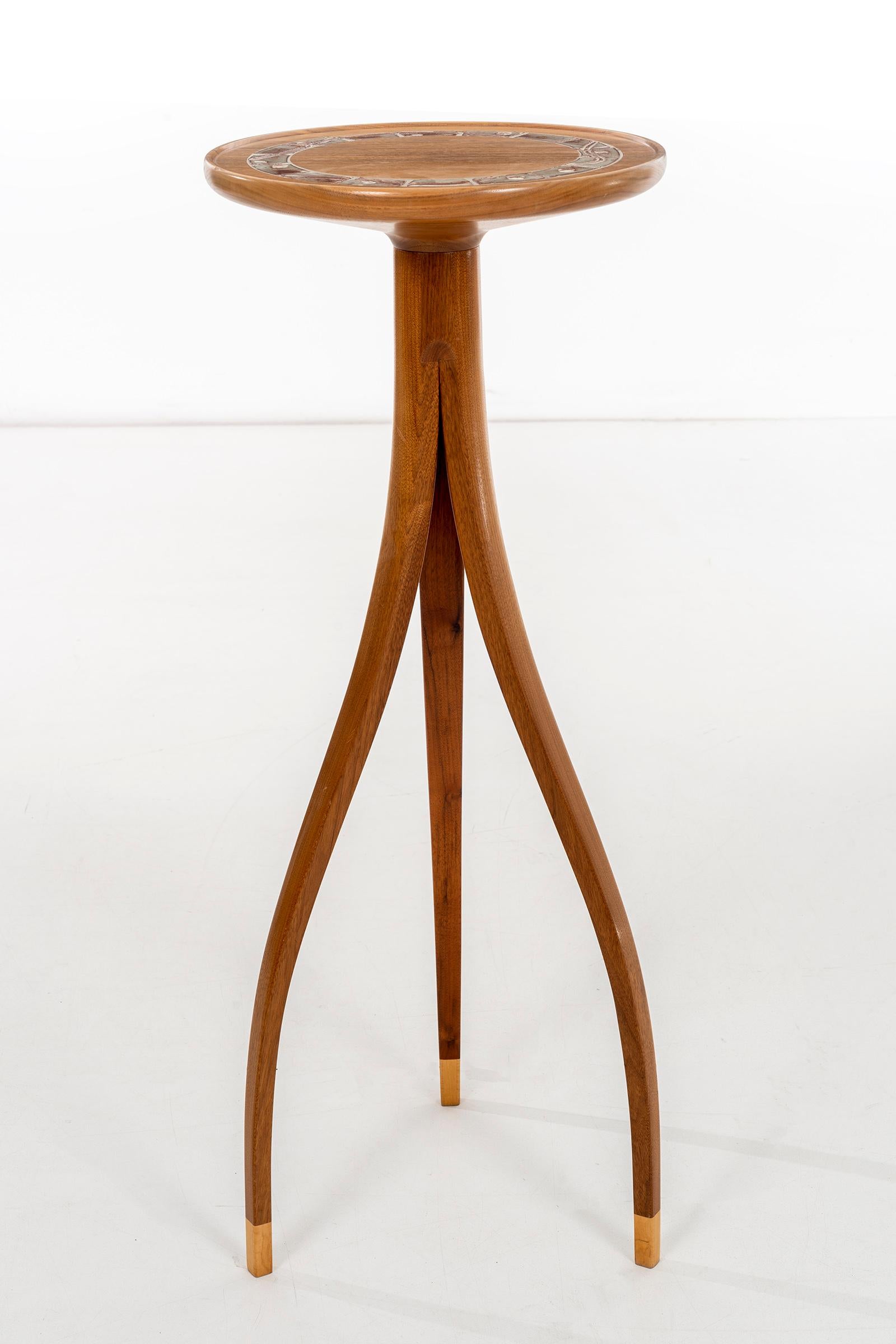 Slender scale high tripod table, solid carved walnut lip edge top with ceramic design detail inset.
Feet have lighter tone wood accent.