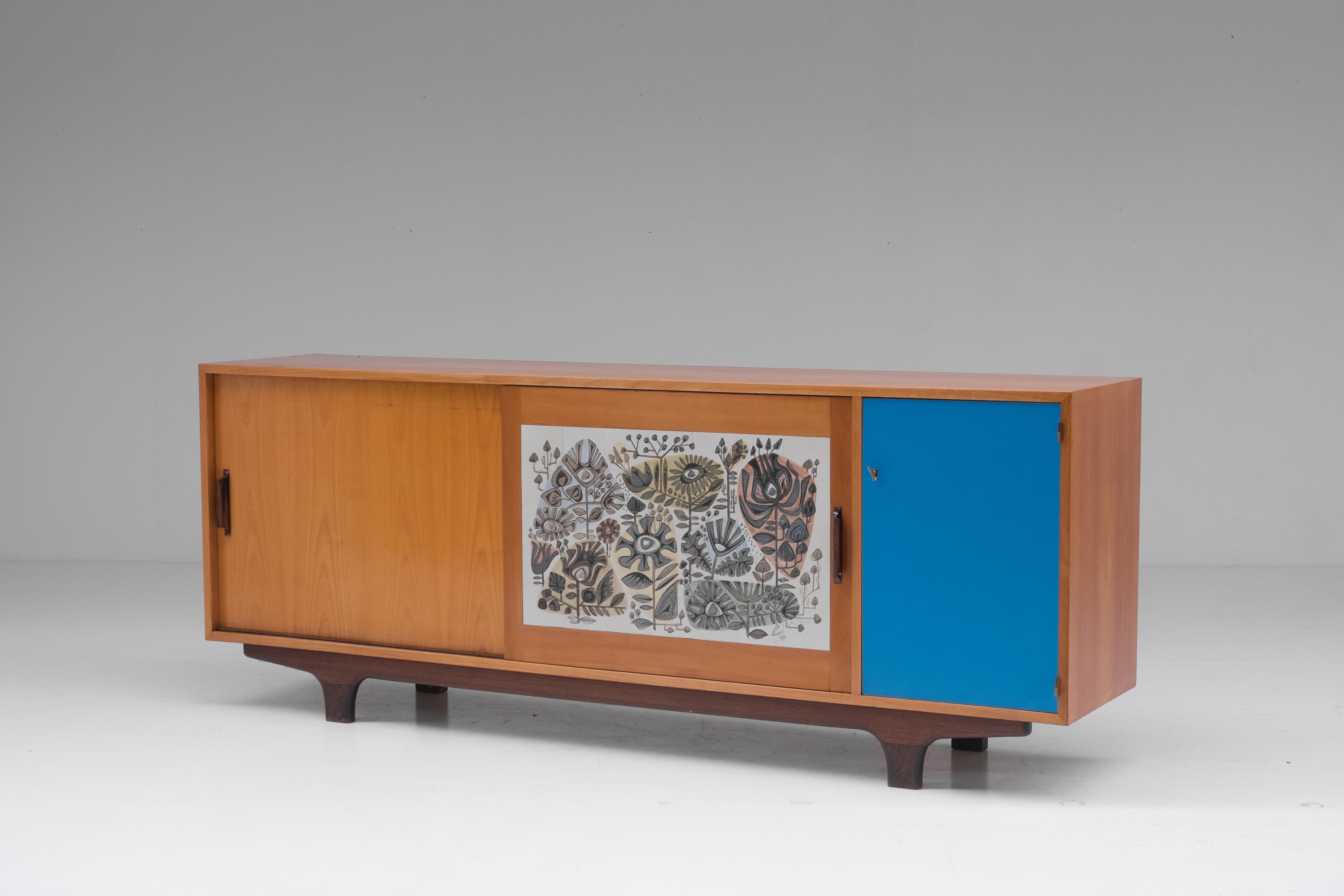 Midcentury modernist credenza with sliding doors from the 1950s, Belgium.
The sideboard shows some very prolific details which shows signs of great quality.
Just look at the custom designed ceramic Perignem piece on the door panel, the sky blue