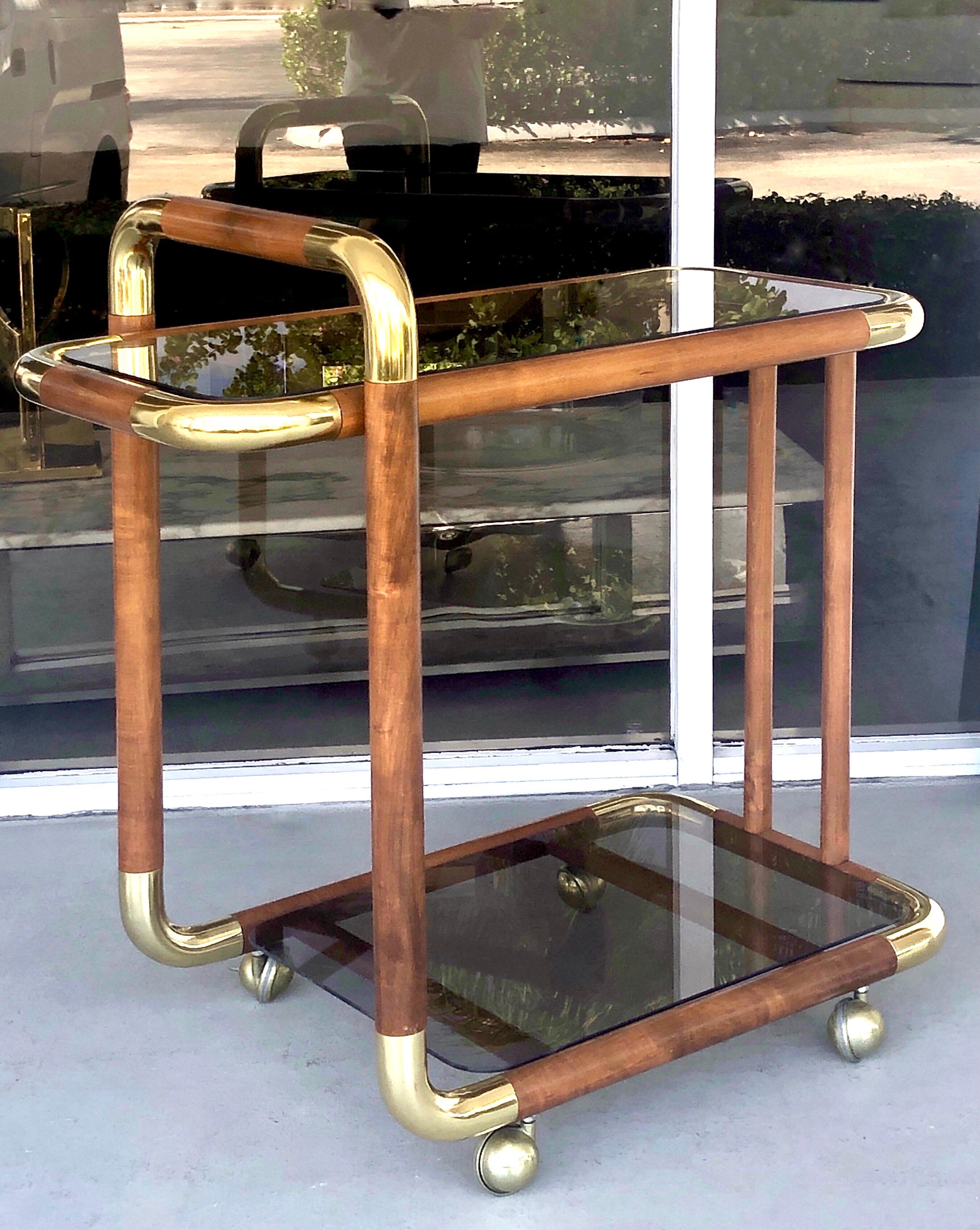 A 1970s bar cart. A thick solid wood frame with radial brass corners. Bronze color glass surfaces. Quite unique with wonderful lines and great looking.