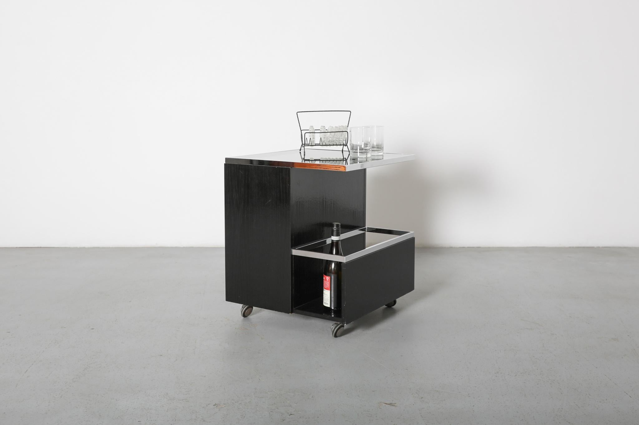 Un elegante carrello da bar nero in stile Bauhaus, definito da linee pulite e alta funzionalità. Questo carrello a rotelle bifacciale presenta un sofisticato piano in vetro grigio fumé che aggiunge un tocco di eleganza alla sua forma minimalista e