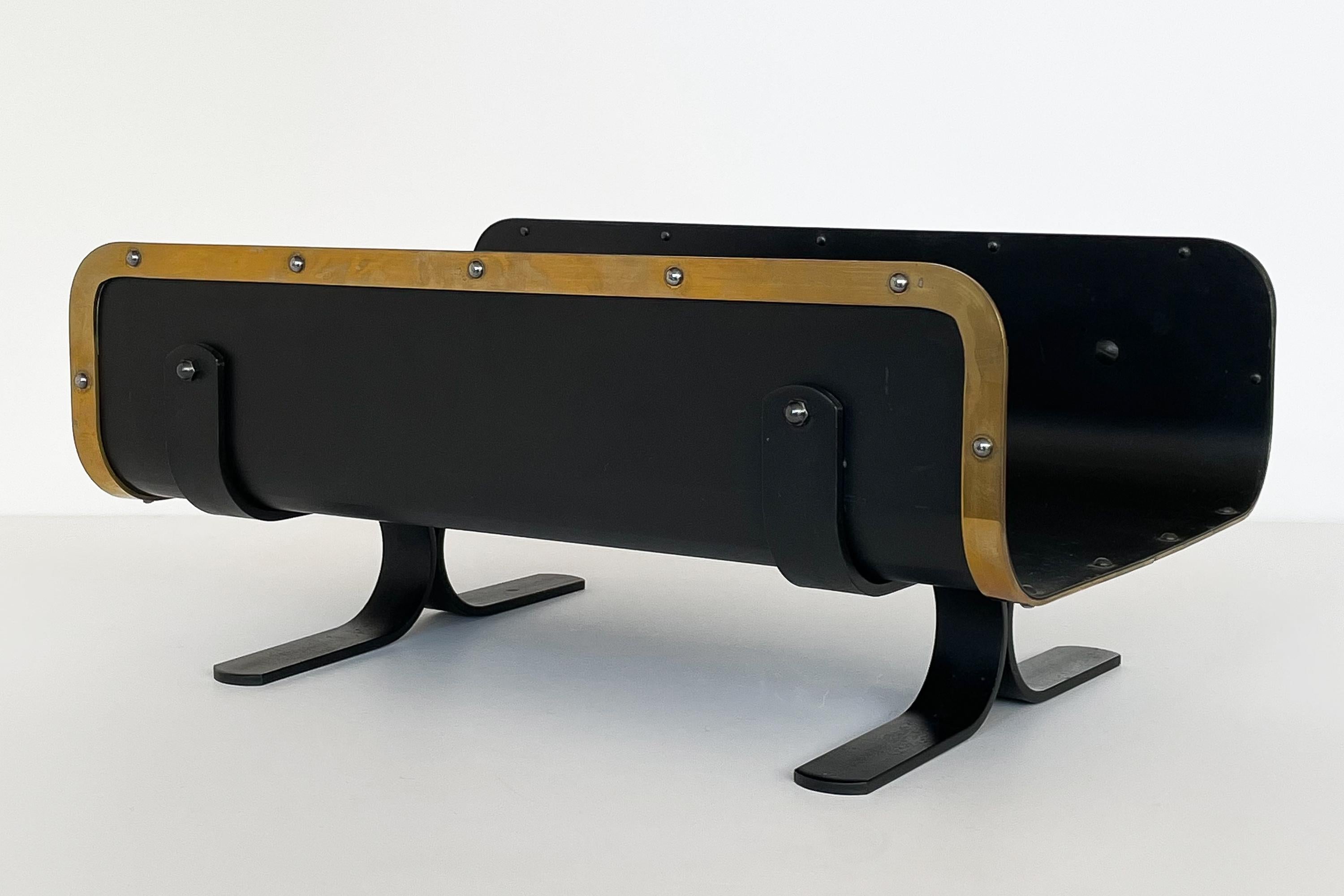 Modernist / mid century log holder with riveted brass edge trim, in the style of Donald Deskey, circa 1960s. Consider using as a magazine rack. Blackened steel and brass with splayed pedestal legs. General patina to brass and slight wear to black