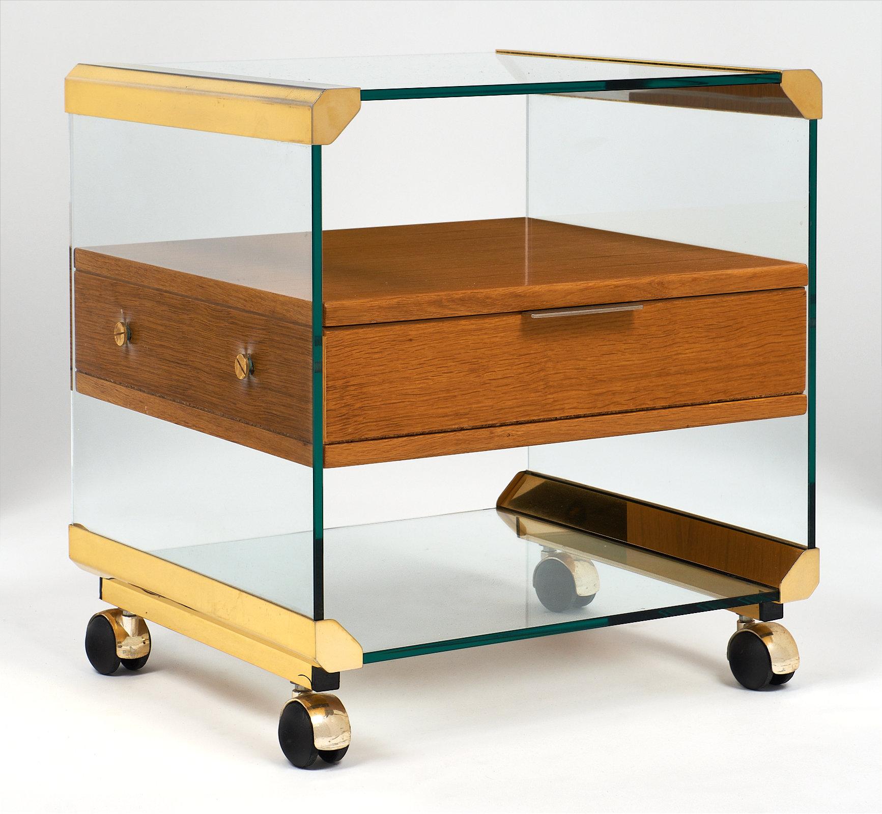 A fun brass and glass modernist side table or nightstand featuring one dovetailed drawer. There is a glass structure encasing the rosewood drawer cabinetry, and brass hardware though out. Four casters support this great piece!