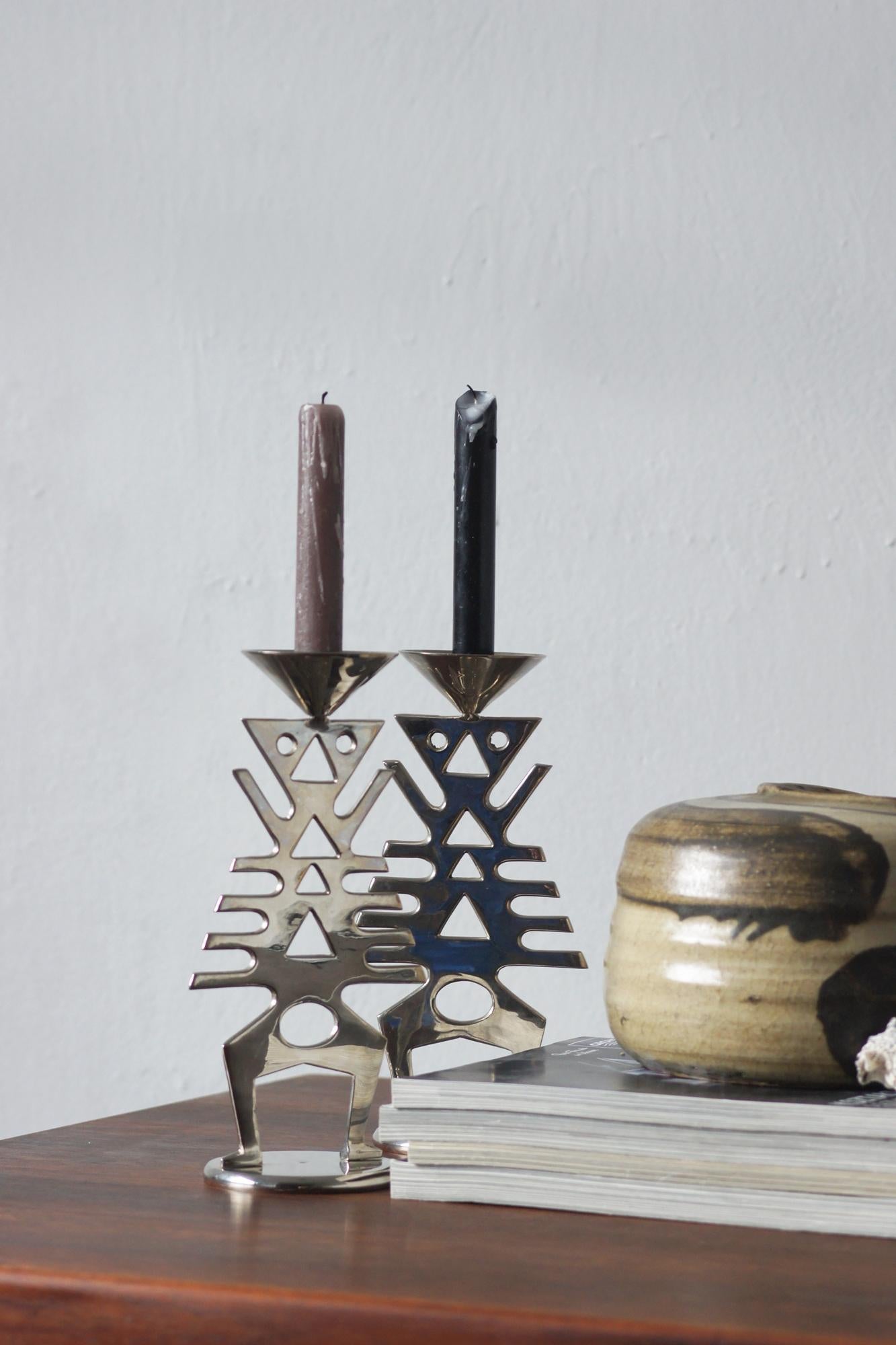 A set of two candle holders made of metal with a polished surface, designed in a modernist style. Their form is inspired by geometric and abstract motifs – cut-out shapes of triangles, circles, and straight lines form an almost totemic silhouette,