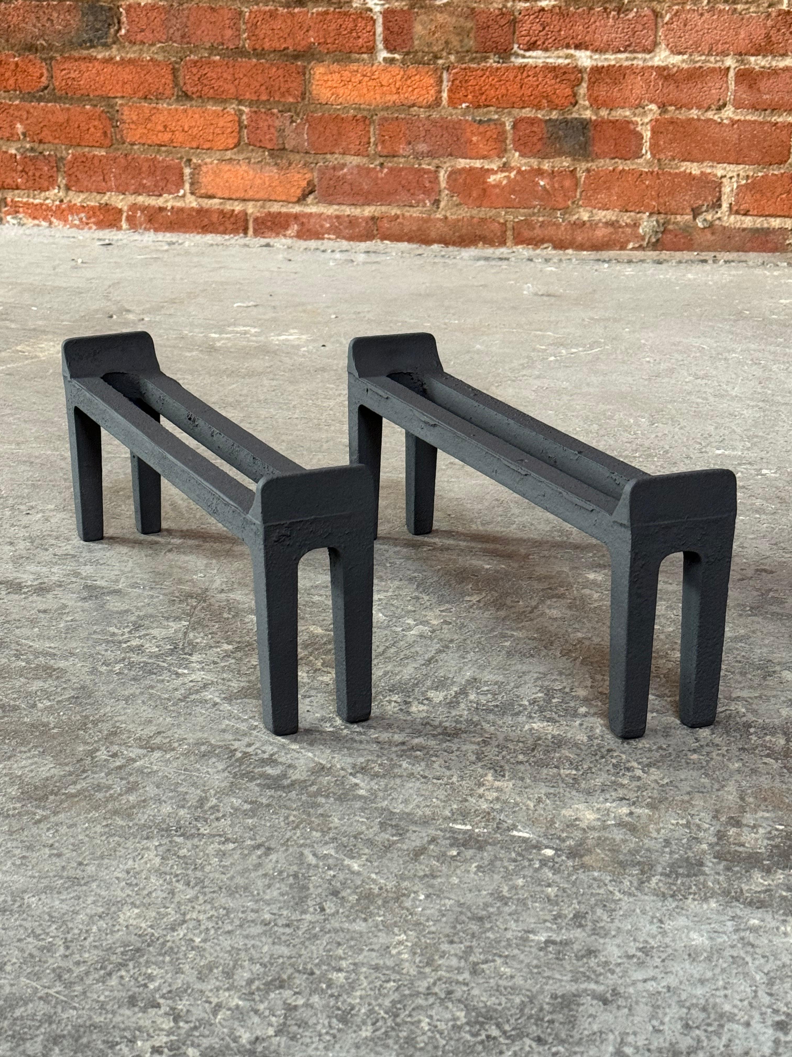 Modernist design andirons by the American company Peerless circa 1950s. A simple minimal design constructed of cast iron and treated with a high temperature rated ceramic based paint in a flat black finish. The design has a parallel bar center with