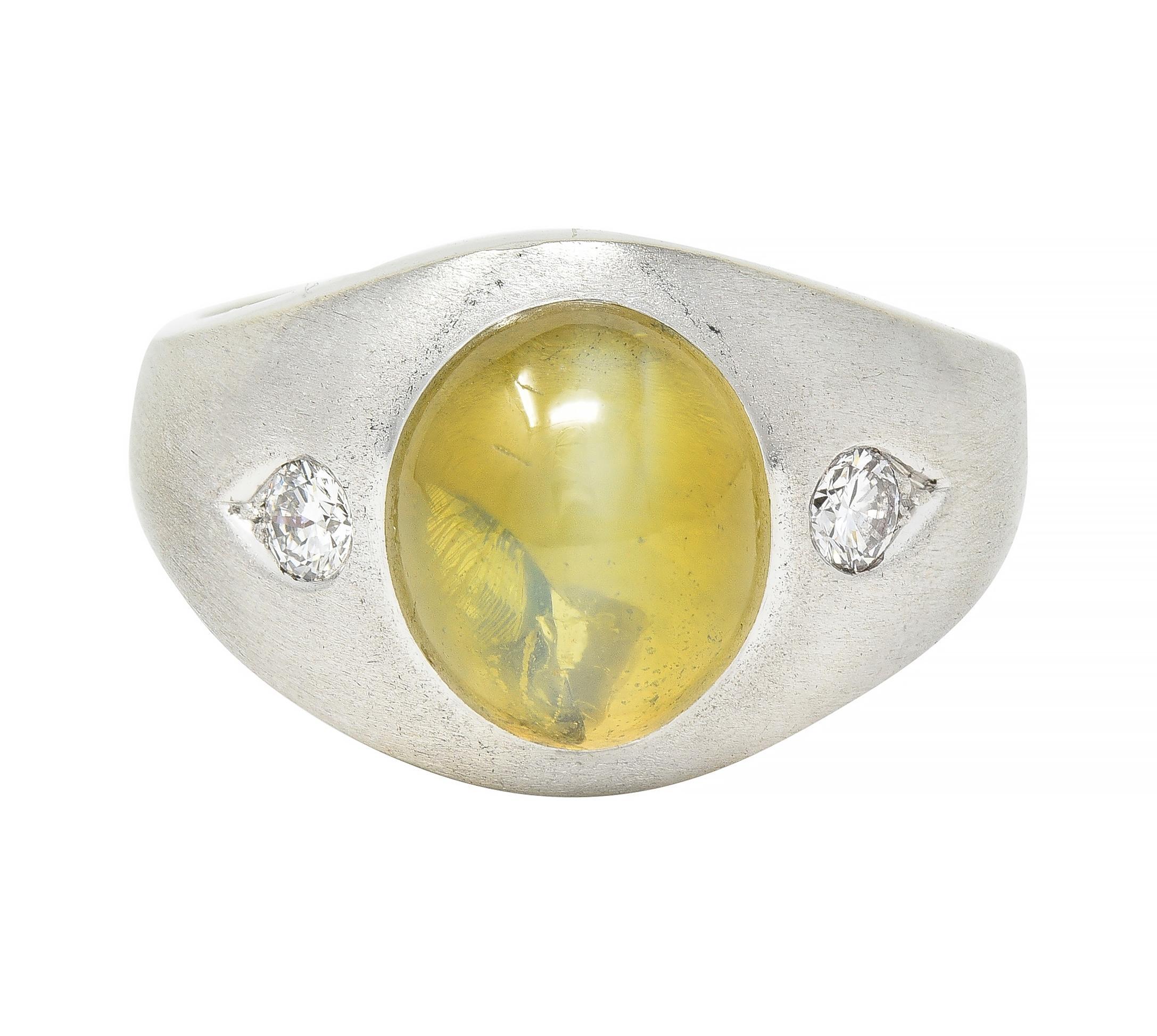 Centering an oval shaped cats eye chrysoberyl measuring 10.9 x 9.3 mm 
Translucent yellowish green displaying linear chatoyancy 
With milk and honey effect and natural mica inclusions 
Flush set in brushed matte gold bombay surround 
Flanked by