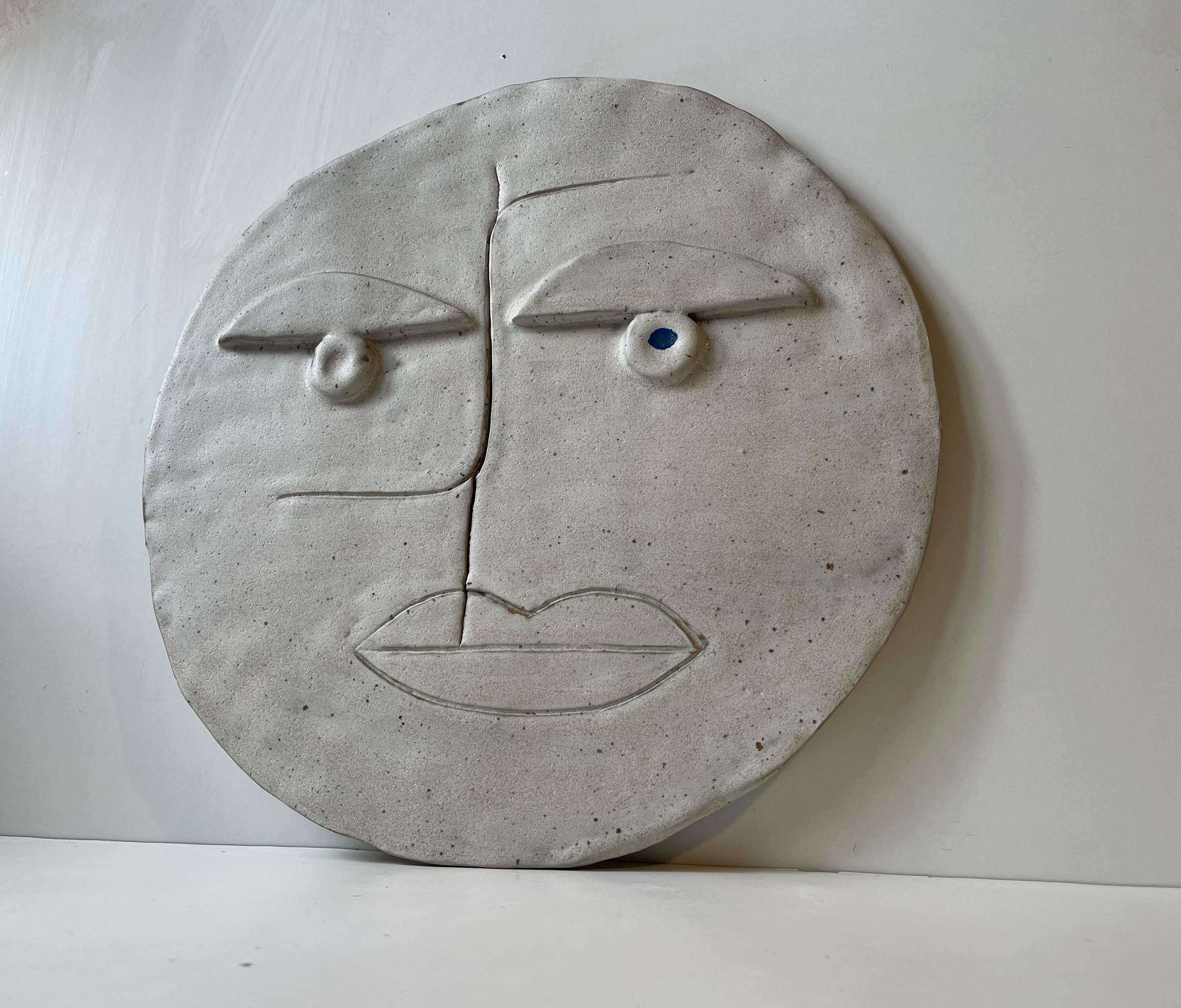 A large circular visage/face wall relief measuring 38x37 cm. It is executed in glazed ceramic and features a blue right eye and an intended crack that runs with the nose down to the mouth. It is made by an unknown/un-identified Scandinavian artist