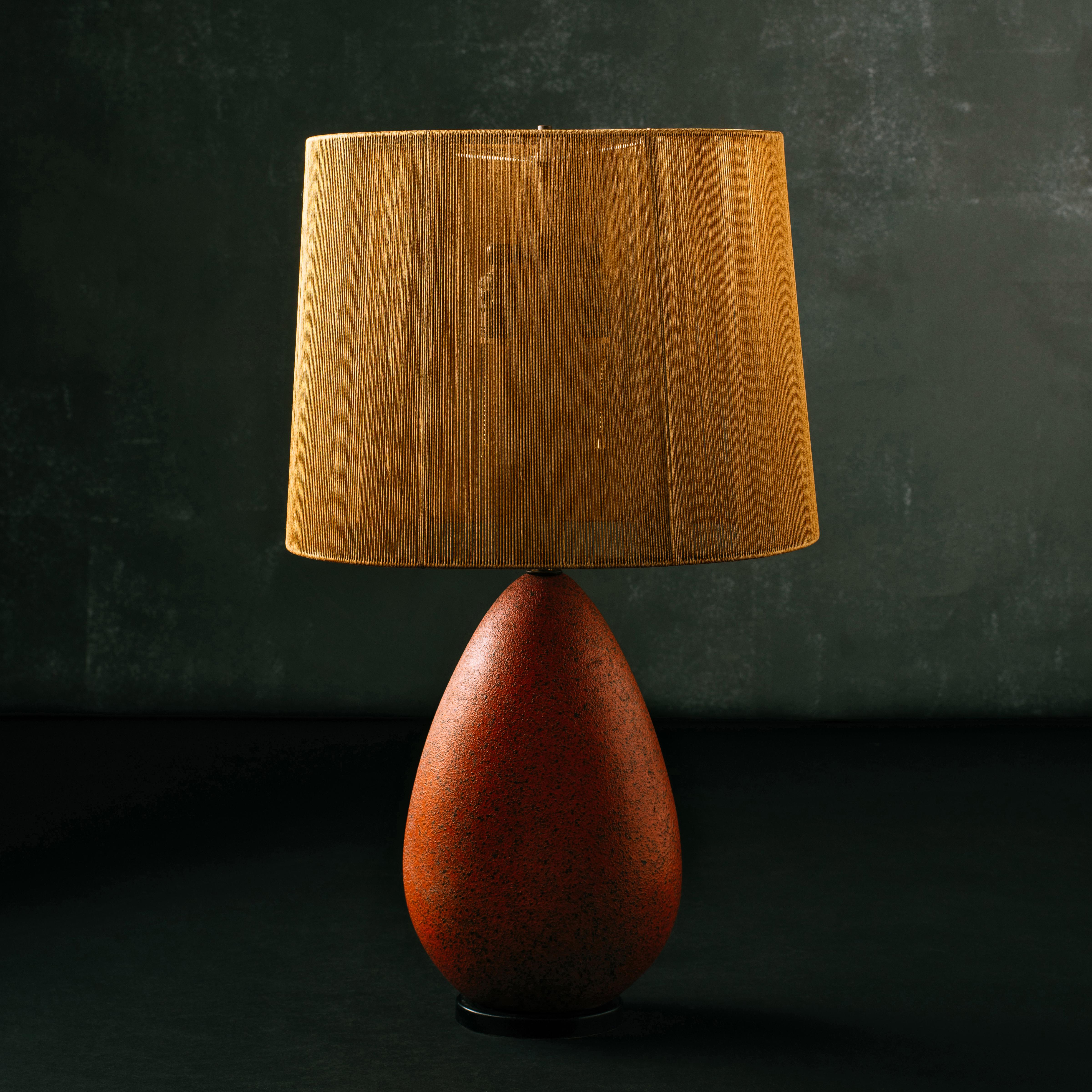 Modern table lamp in an oval teardrop form with a coarse, matte glaze in brick red, supported by an ebonized wood base, and with its rare, original string shade providing warm illumination. Double socket hardware with inline switch to original