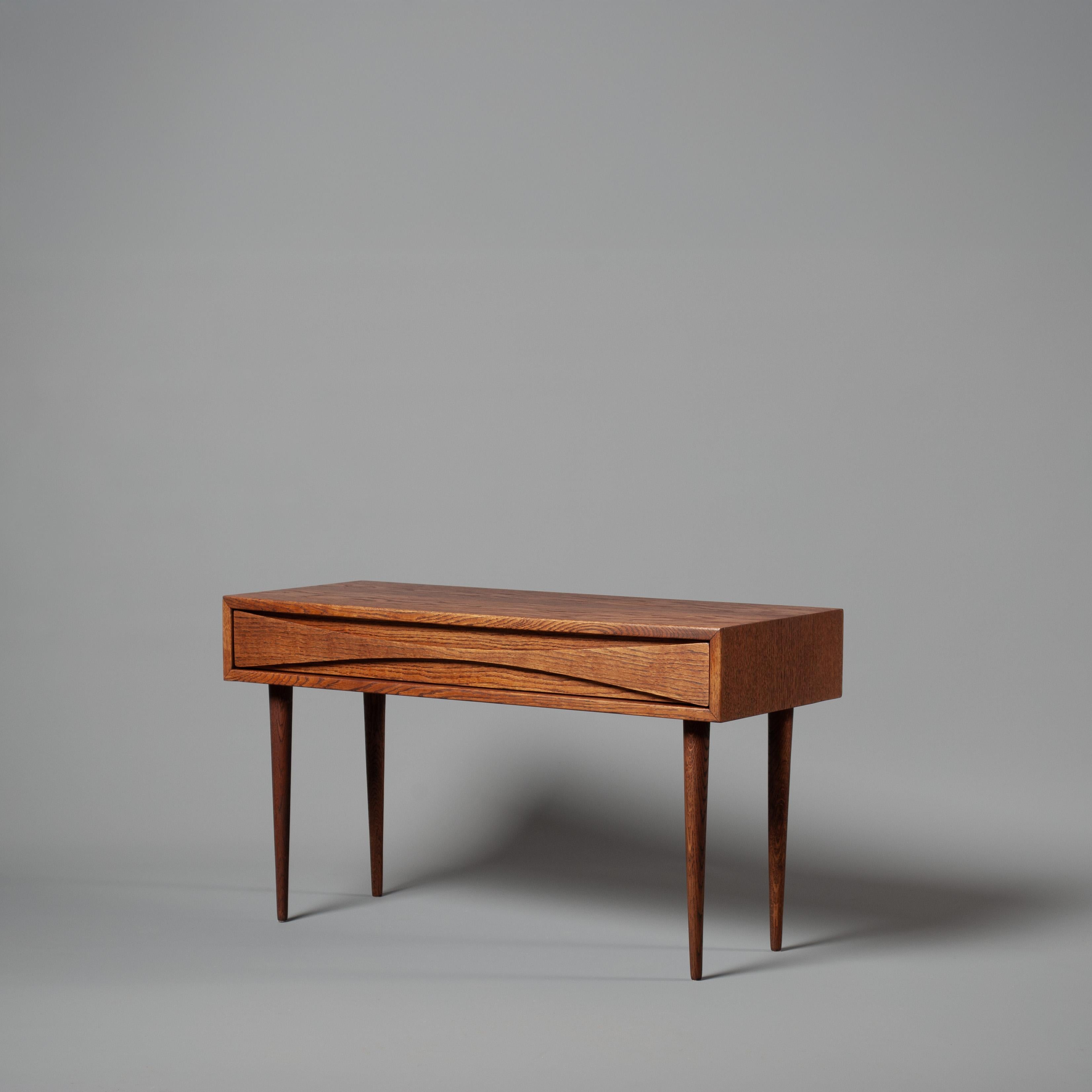 Il raro e bellissimo tavolo a cassetto singolo di Niels Clausen. Esterno in rovere scuro, interno dei cassetti in rovere e maniglia con curva a forma di arco. Gambe affusolate e sottili. Prodotto da NC Mobler, Danimarca, circa 1960. 
Un mobile