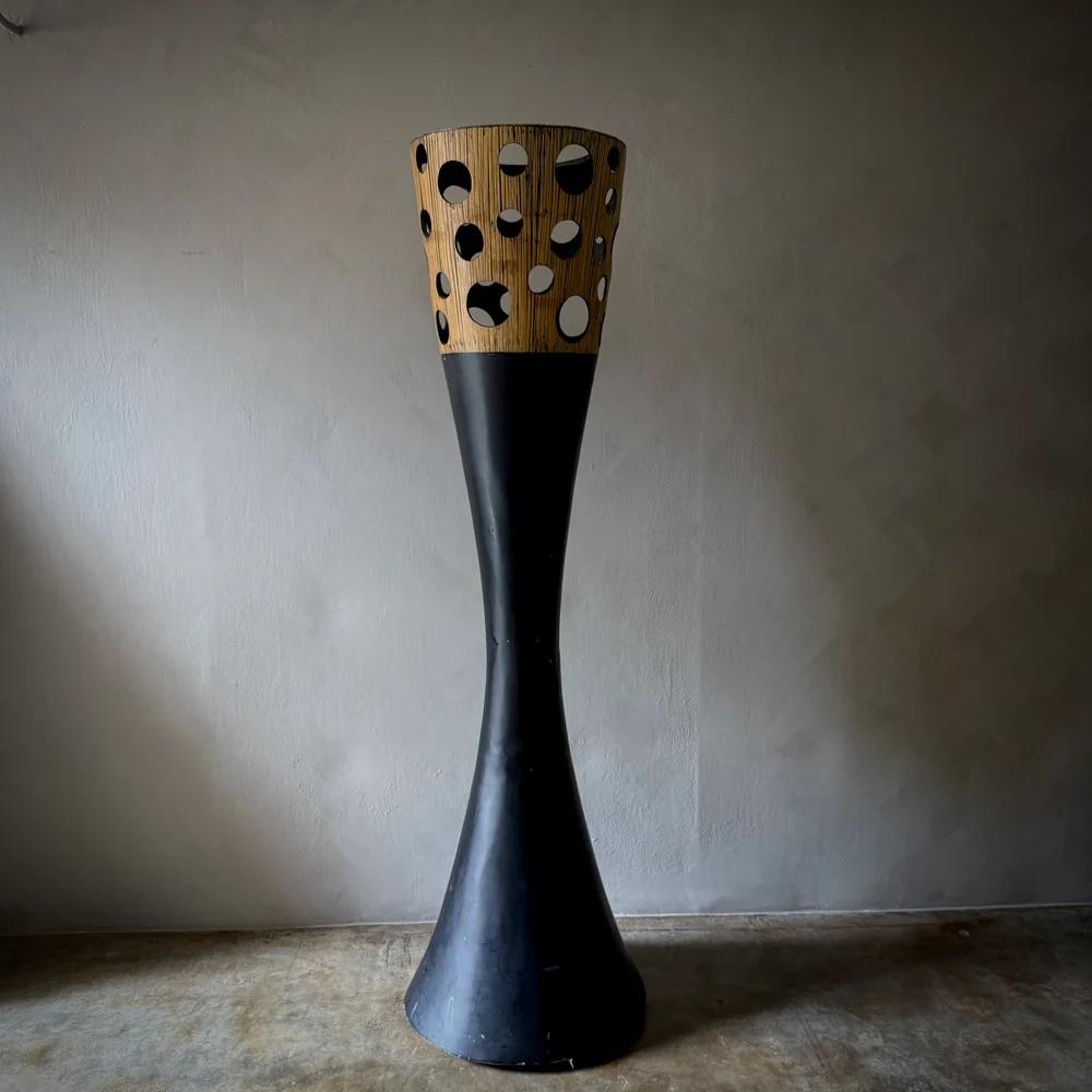 This sculptural column blends bold midcentury design with a futuristic edge, featuring a flared black base that elegantly tapers into a perforated top section of warm-toned wood. The contrasting materials and organic cut-out shapes create a dynamic