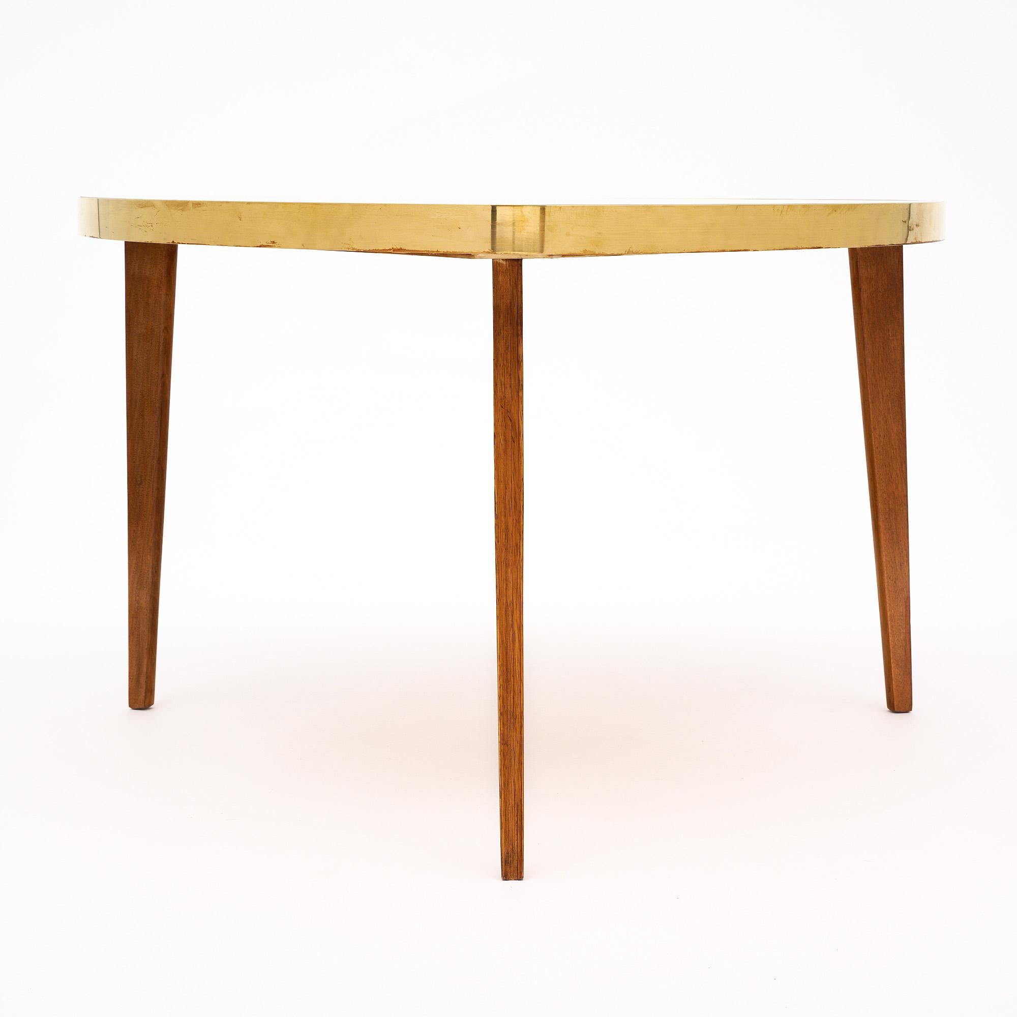 Table basse tripode de France de style moderniste. Cette pièce a des pieds en bois effilés qui soutiennent un plateau avec des garnitures en laiton. La forme triangulaire incurvée est dotée d'un plateau en verre recouvert de parchemin pour un effet