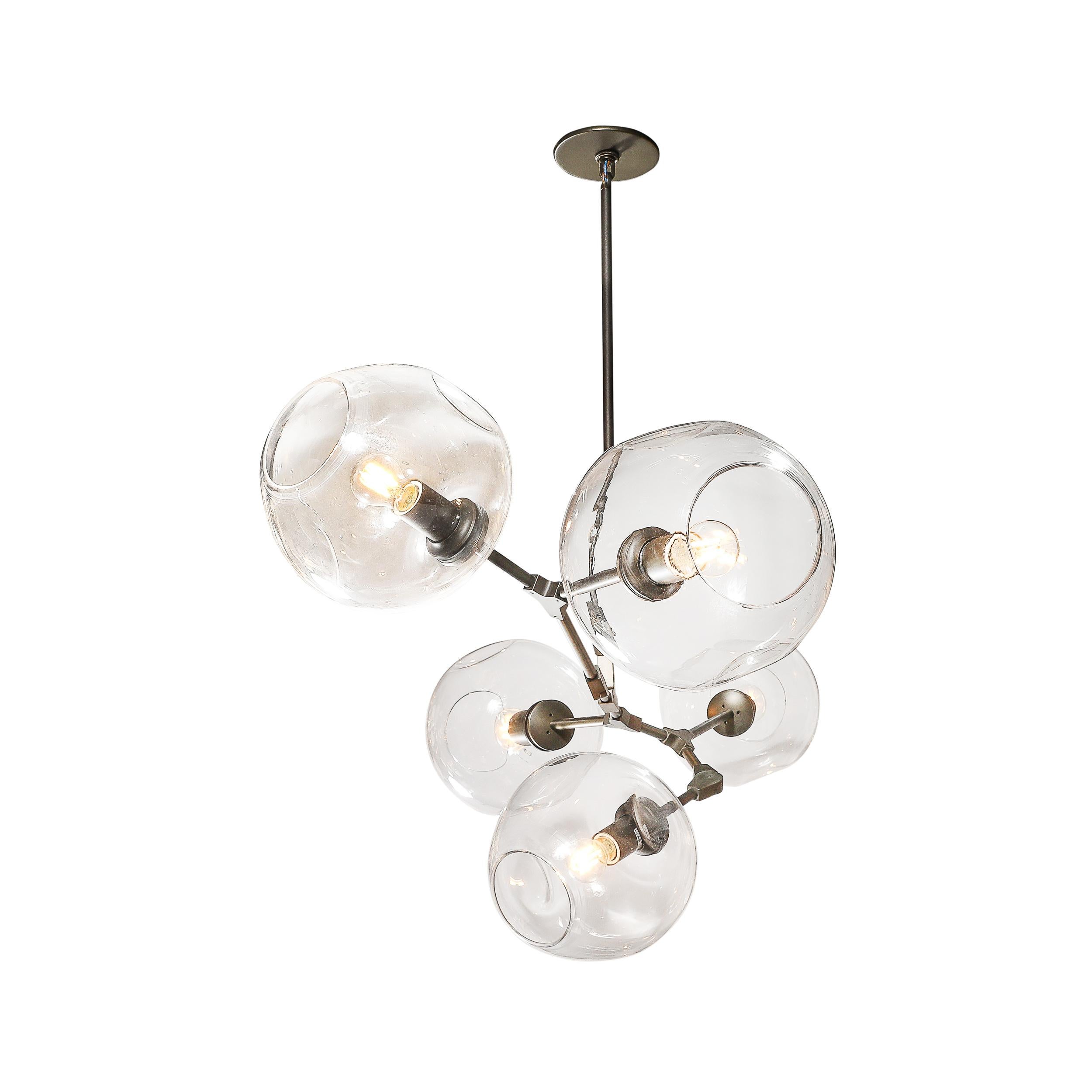 Questo splendido lampadario modernista della celebre designer Lindsey Adelman presenta un'armoniosa miscela di eleganza industriale e artigianalità. Con cinque globi di vetro trasparente soffiato a mano, il suo design evidenzia la bellezza scultorea