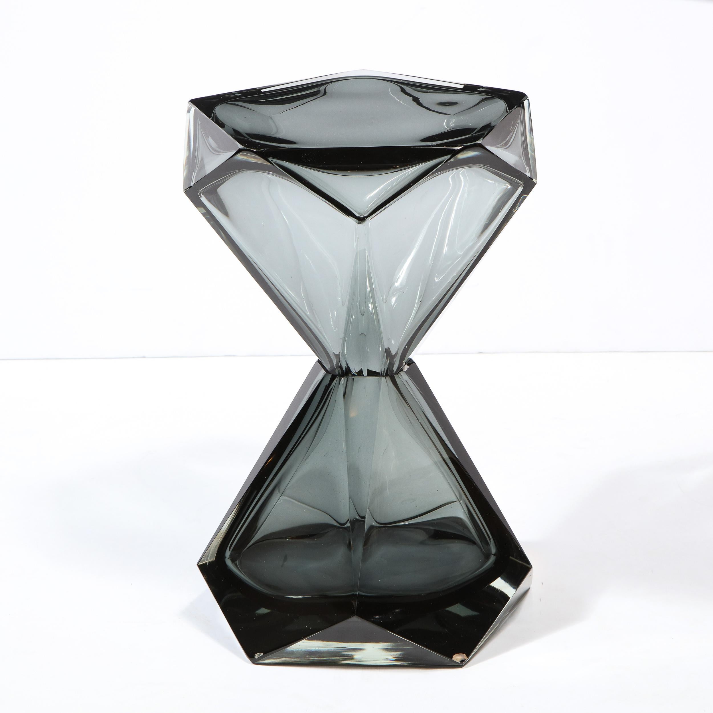 This refined and graphic modernist side/end table was realized in Murano, Italy- the island off the coast of Venice renowned for centuries for its superlative glass production. It features a faceted hourglass form body in smoked translucent graphite