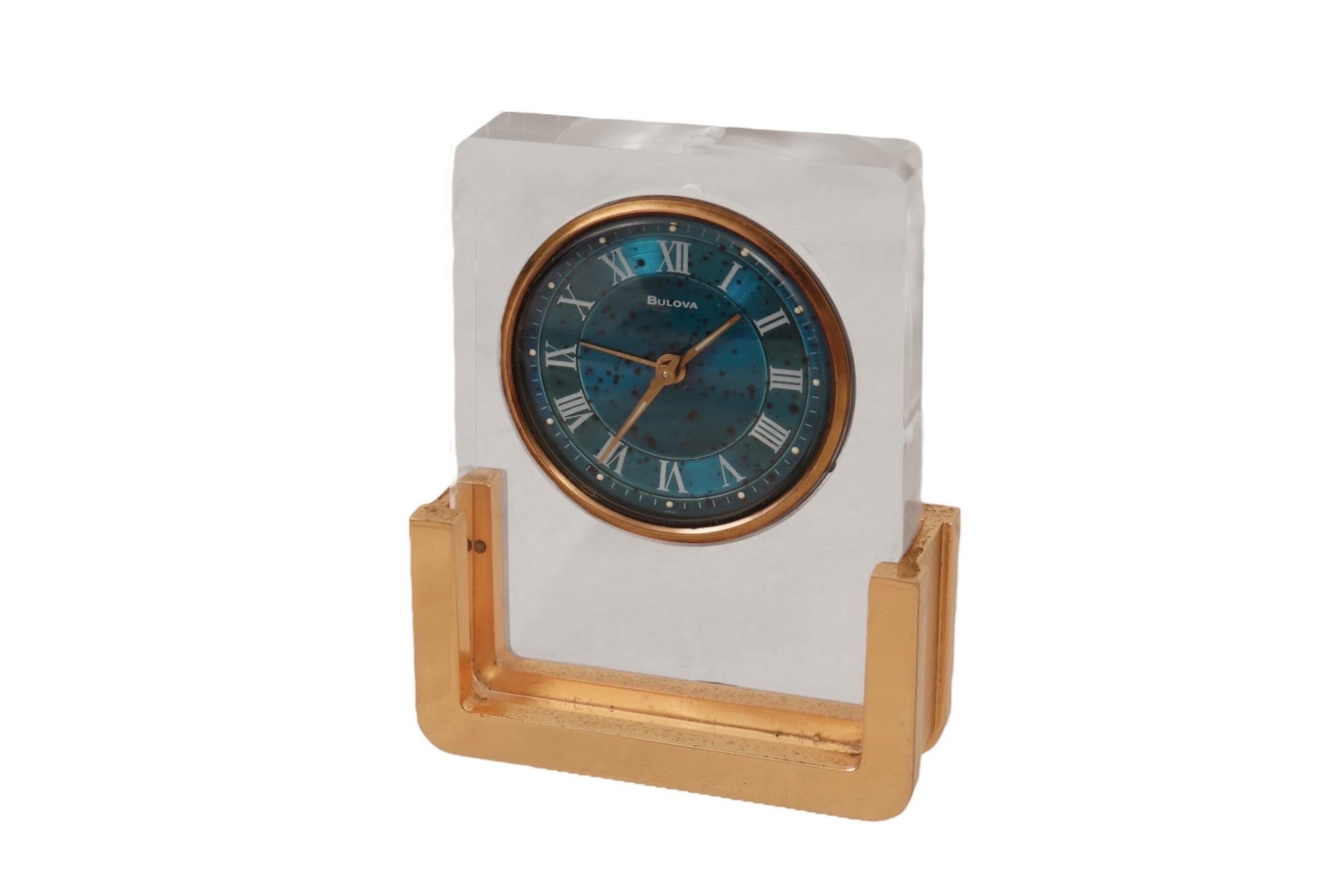 Réveil de table moderniste modèle 2RA007 fabriqué par Bulova au Japon. Un corps en Lucite transparent renferme le cadran bleu de l'horloge et le mécanisme de remontage, le tout monté sur une élégante base métallique en laiton.

