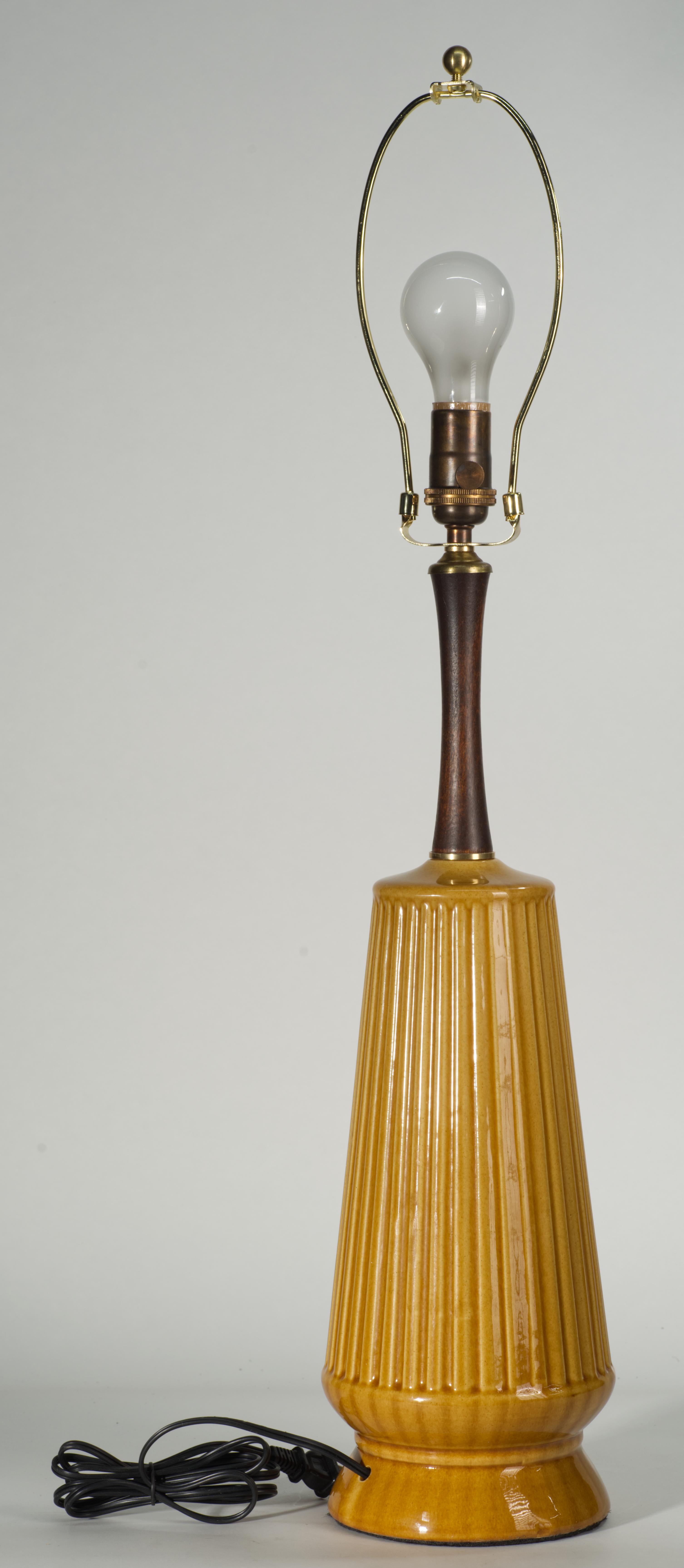  La lámpara de sobremesa alta vintage de cerámica y balaustre de madera presenta un cuerpo alargado de cerámica con un dibujo geométrico texturizado y una base redonda definida, rematada con el cuello de madera tallada. La textura lineal,